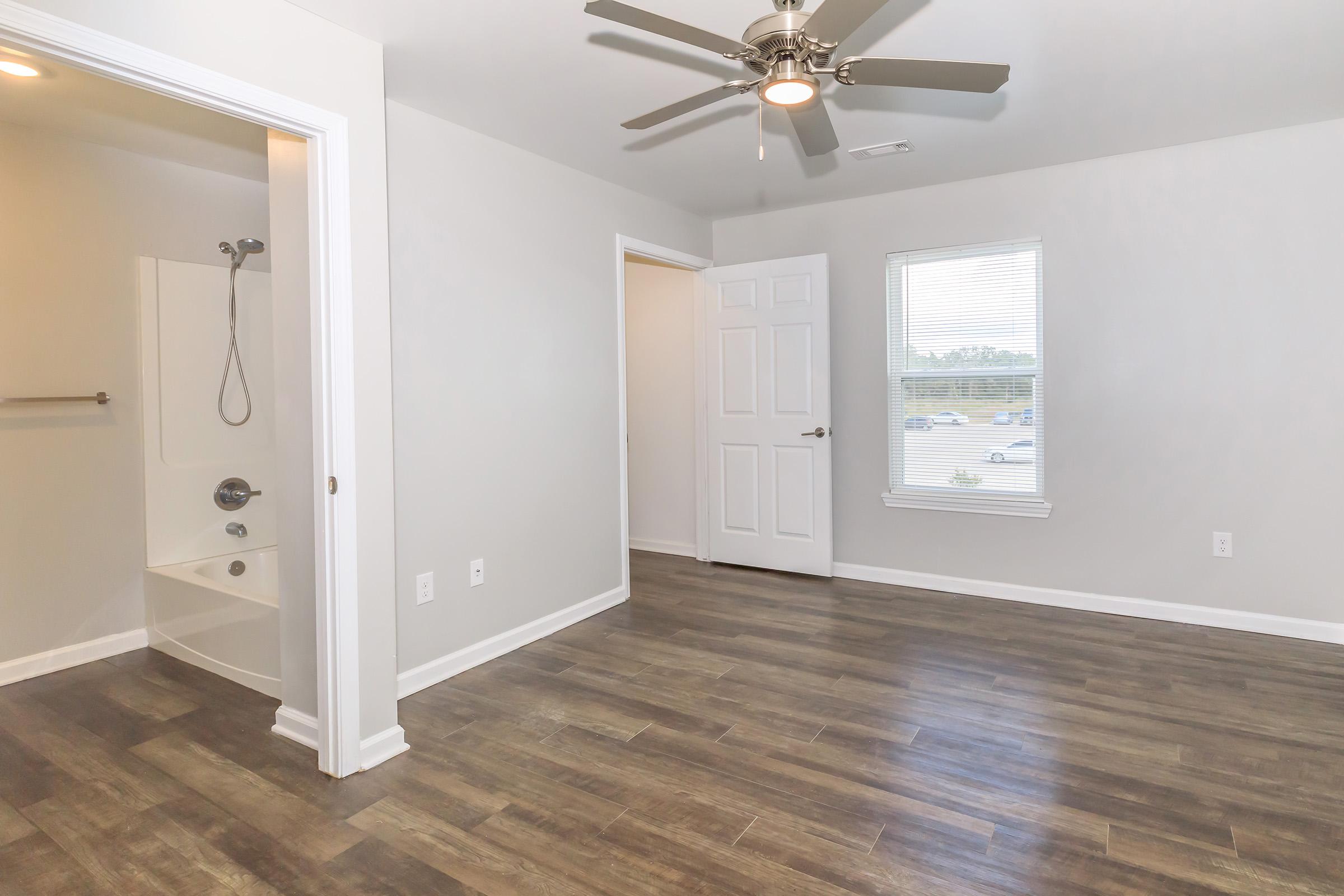 A spacious, modern bedroom with a ceiling fan, light gray walls, and laminate flooring. There is a doorway leading to a small bathroom area with a shower and tub, and a window providing natural light. The overall ambiance is clean and inviting, perfect for comfortable living.