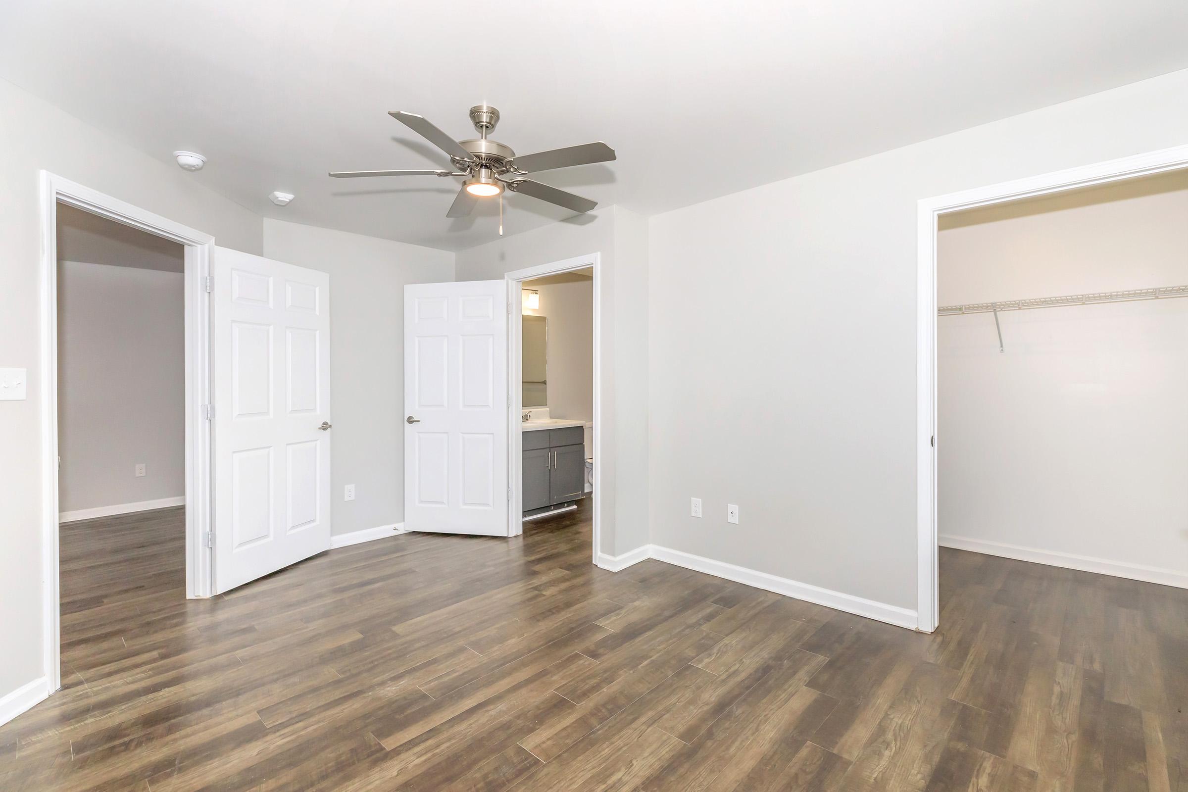 A spacious room featuring light-colored walls, a ceiling fan, and wooden flooring. There are two open doors leading to separate areas, one of which appears to be a closet. The room is well-lit, emphasizing its clean and modern design.