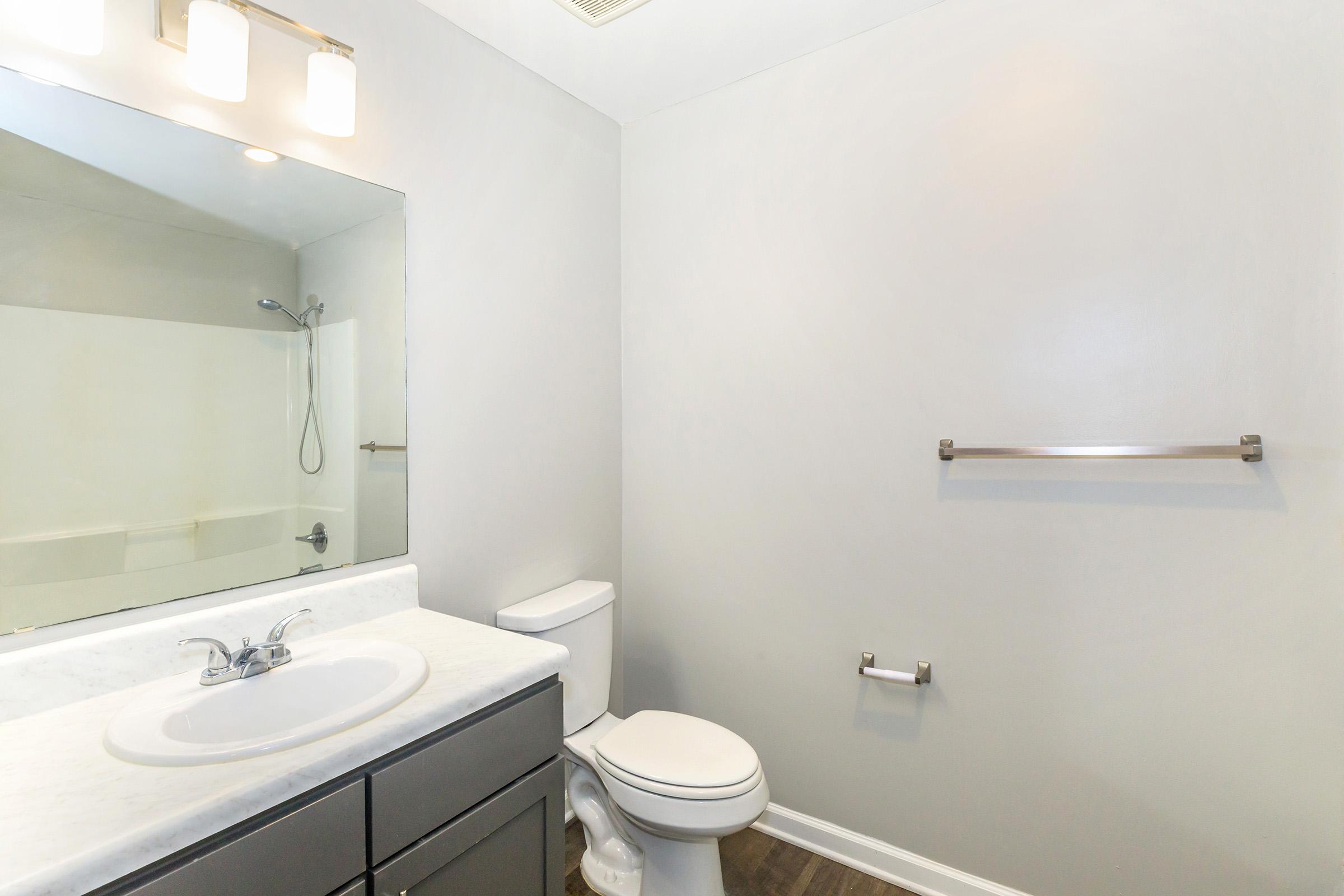 A modern bathroom featuring a white countertop sink with a mirror above, a toilet, and a shower area. The walls are painted light gray, and there is a towel bar next to the toilet. The flooring is dark, adding a contrast to the overall bright aesthetic of the space.