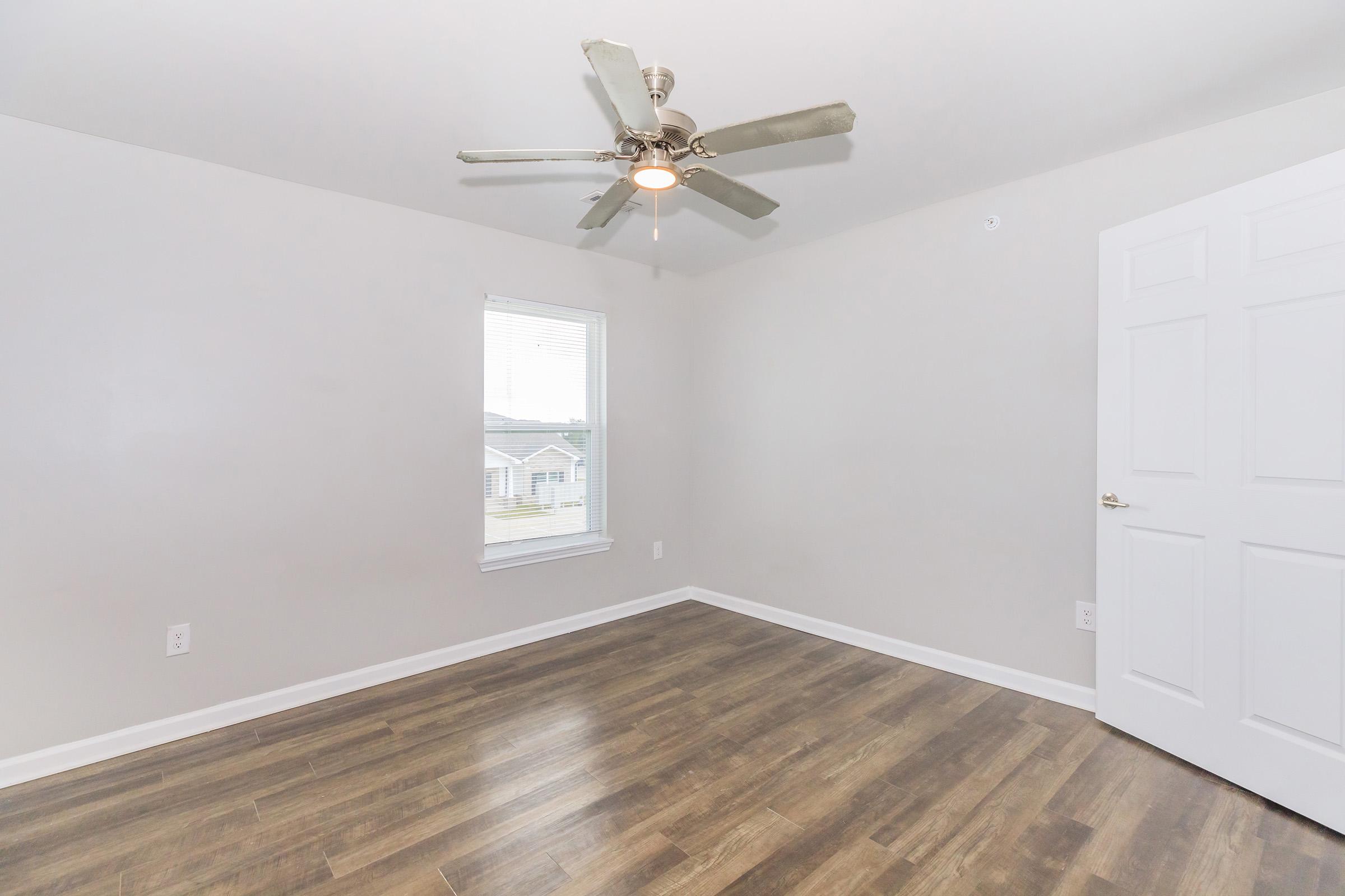 A well-lit empty room with a light-colored wall and a ceiling fan. It features a window with a view outside and a door on the right. The floor is covered with dark wood-like laminate, creating a warm ambiance. Ideal for a bedroom or small office space.