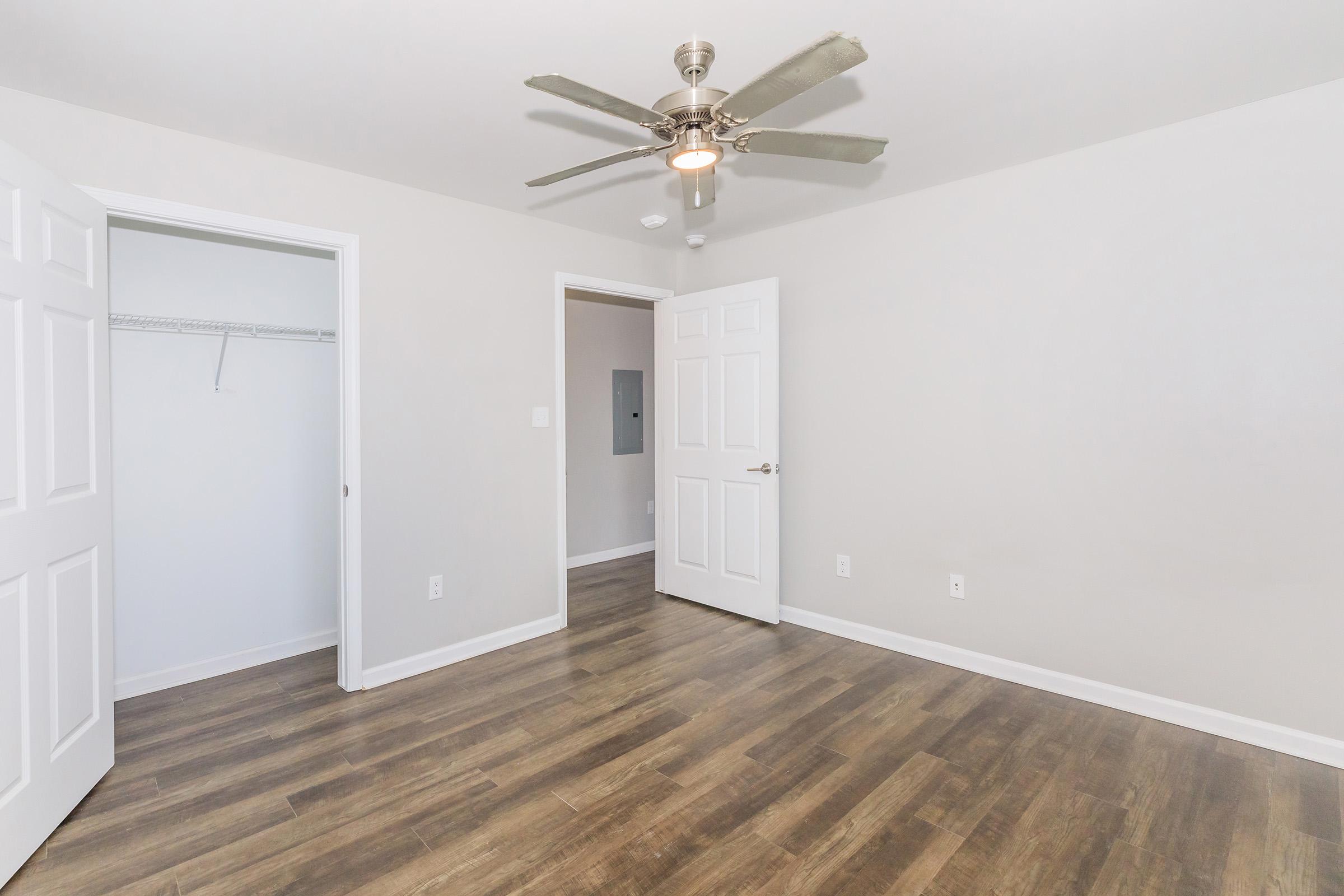 A spacious room featuring a ceiling fan, light-colored walls, and wooden flooring. Two doors lead to different spaces, with one door opening to a closet. The room is well-lit and empty, showcasing a clean and modern interior design.