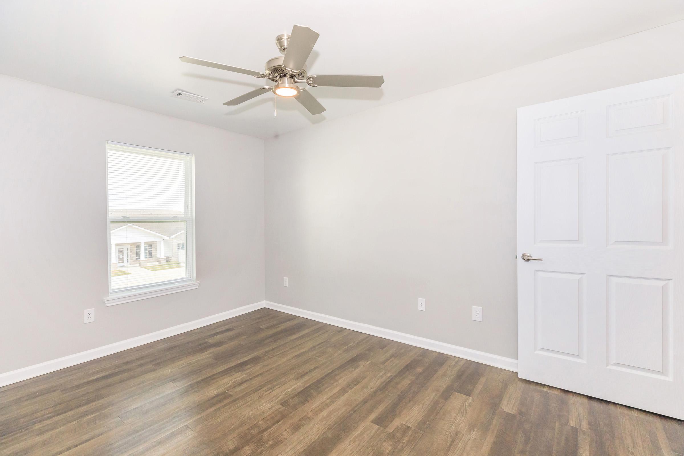A bright, empty room with light gray walls and a ceiling fan. There is a large window letting in natural light, showcasing a view of an exterior area. The floor features dark wooden laminate, and a white door is visible on the right side of the image. The overall space appears clean and modern.
