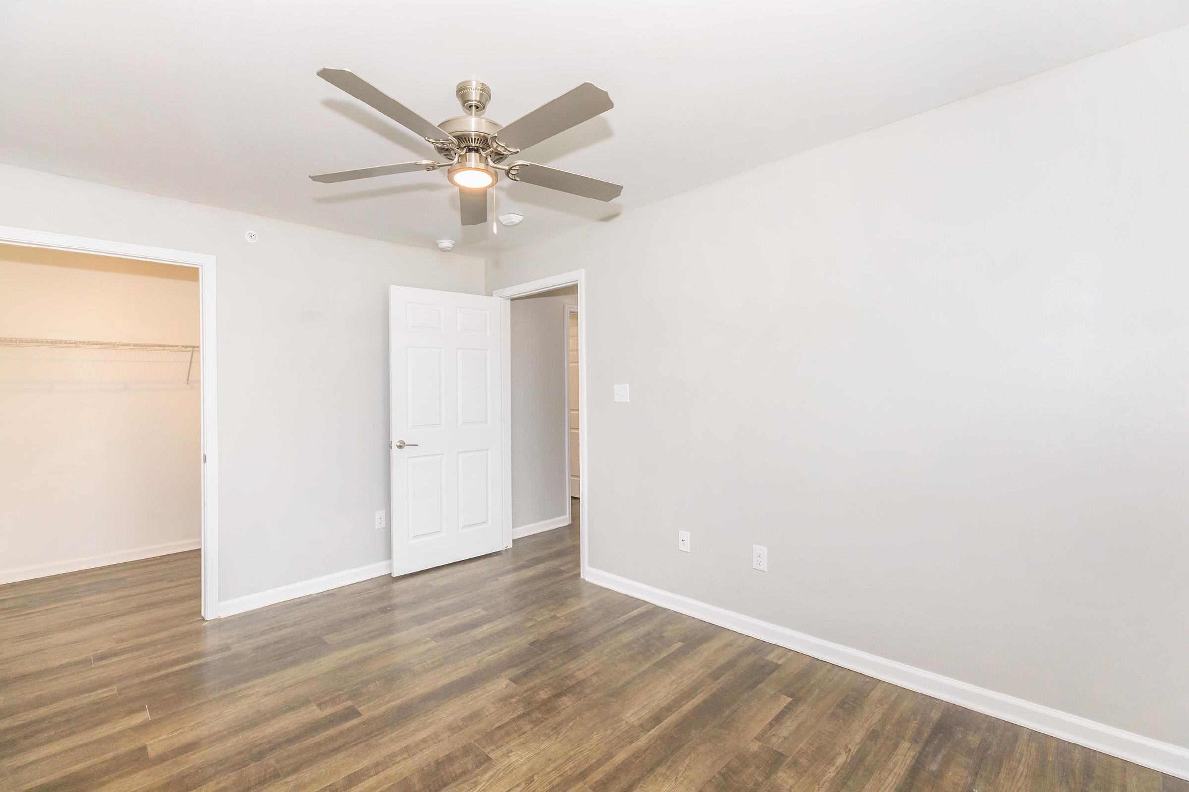 A well-lit room with light gray walls, hardwood flooring, and a ceiling fan. There are two doorways: one leading to a closet and another to an adjoining room. The space appears empty and ready for furnishing, creating a clean and airy atmosphere.