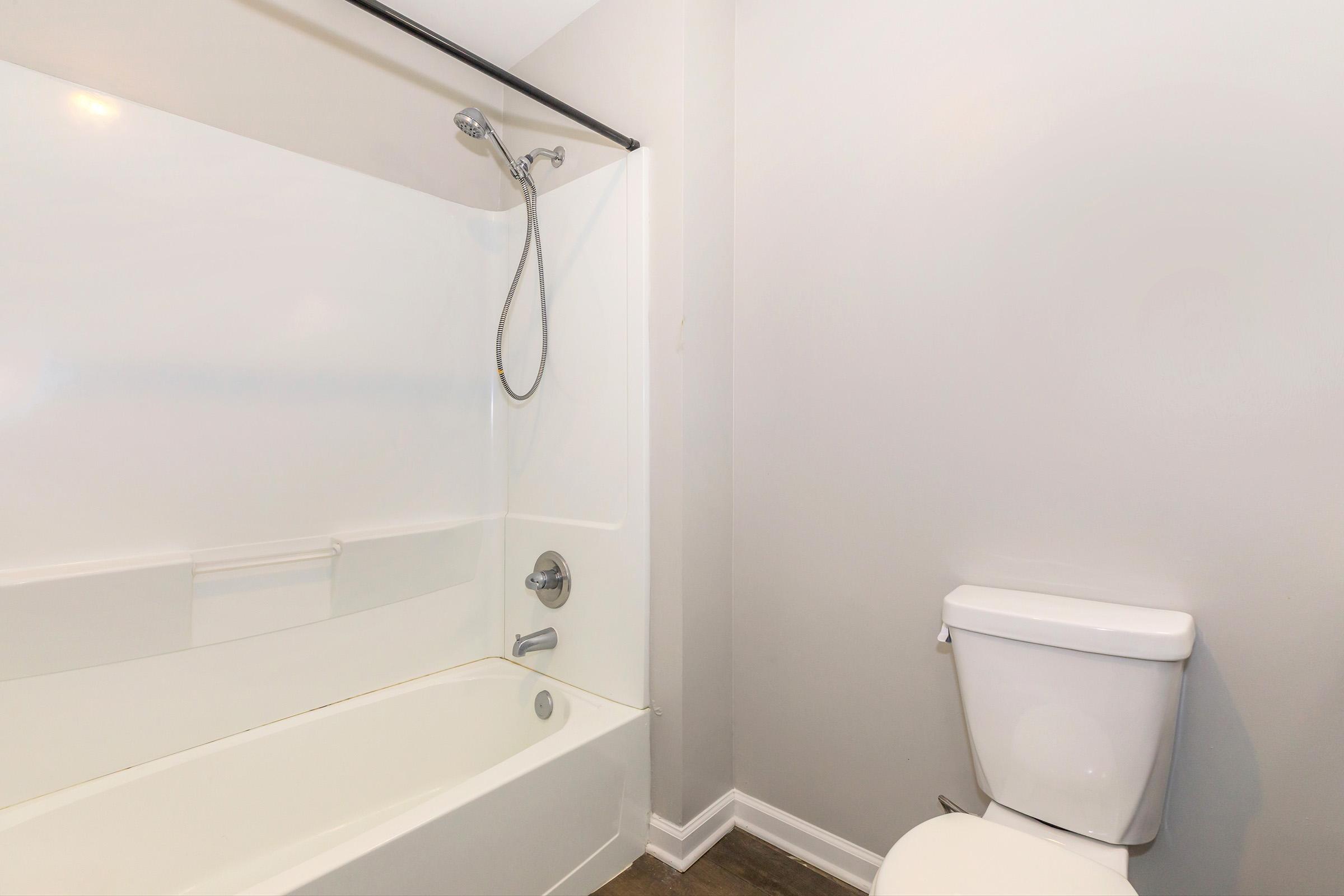 A clean, modern bathroom featuring a white bathtub with a showerhead attached, a toilet beside it, and light gray walls. The space has a minimalist design with no visible decorations or clutter. The flooring is a neutral color, enhancing the bathroom's simplicity and functionality.