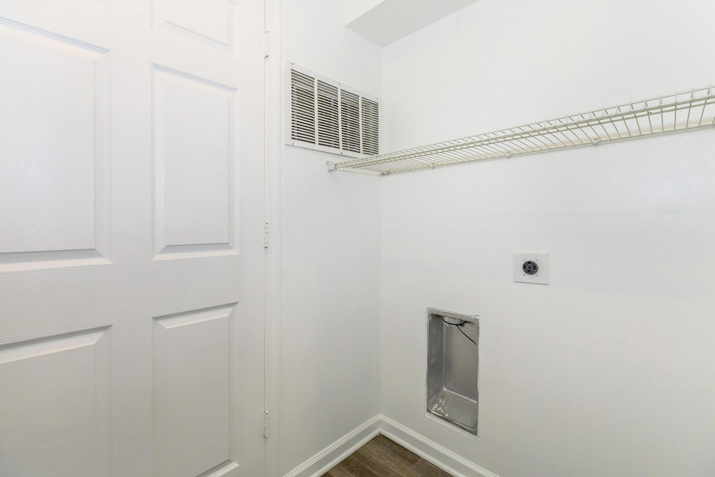 A clean, white-walled closet featuring a ventilated door, a wire shelf along one wall, and an access panel on the lower area. The floor is a light-colored laminate, and the closet has a simple, uncluttered design suitable for storage.