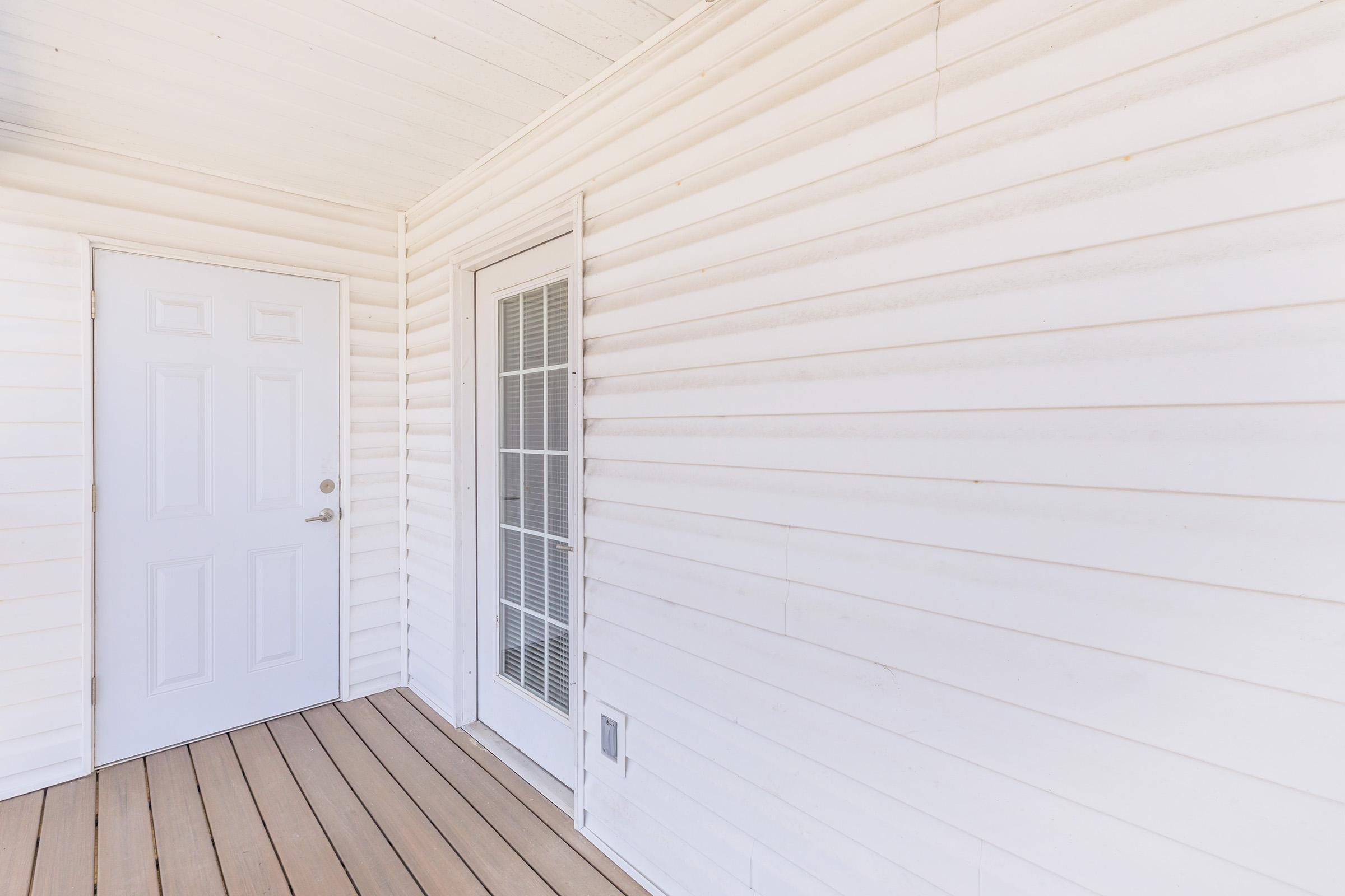 A white exterior wall with horizontal siding, featuring a closed white door and a sliding door with multiple glass panes. The flooring is made of light-colored wooden planks, and the area appears well-lit and uncluttered.