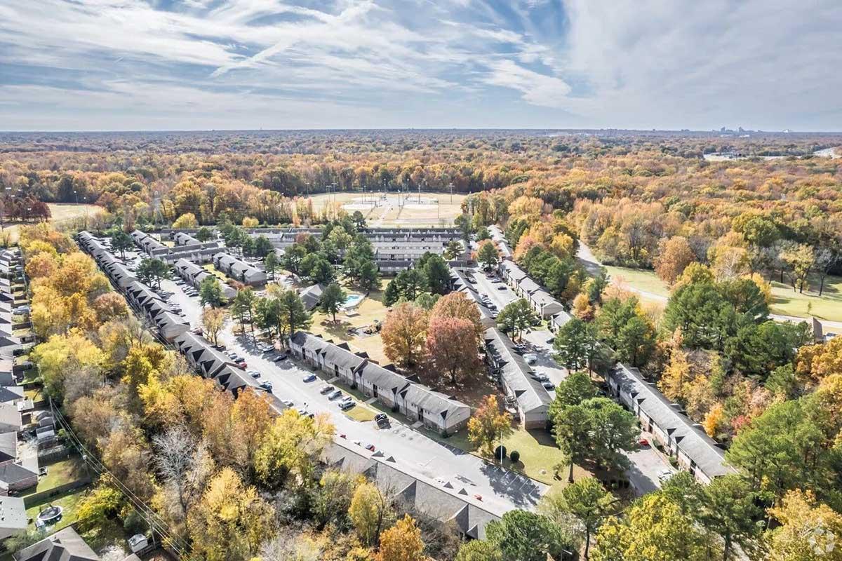 Aerial view of a residential area with low-rise buildings surrounded by trees showing fall colors. In the background, a baseball field is visible, and the landscape features a mix of green and orange foliage, indicating autumn. The scene captures a peaceful suburban environment.