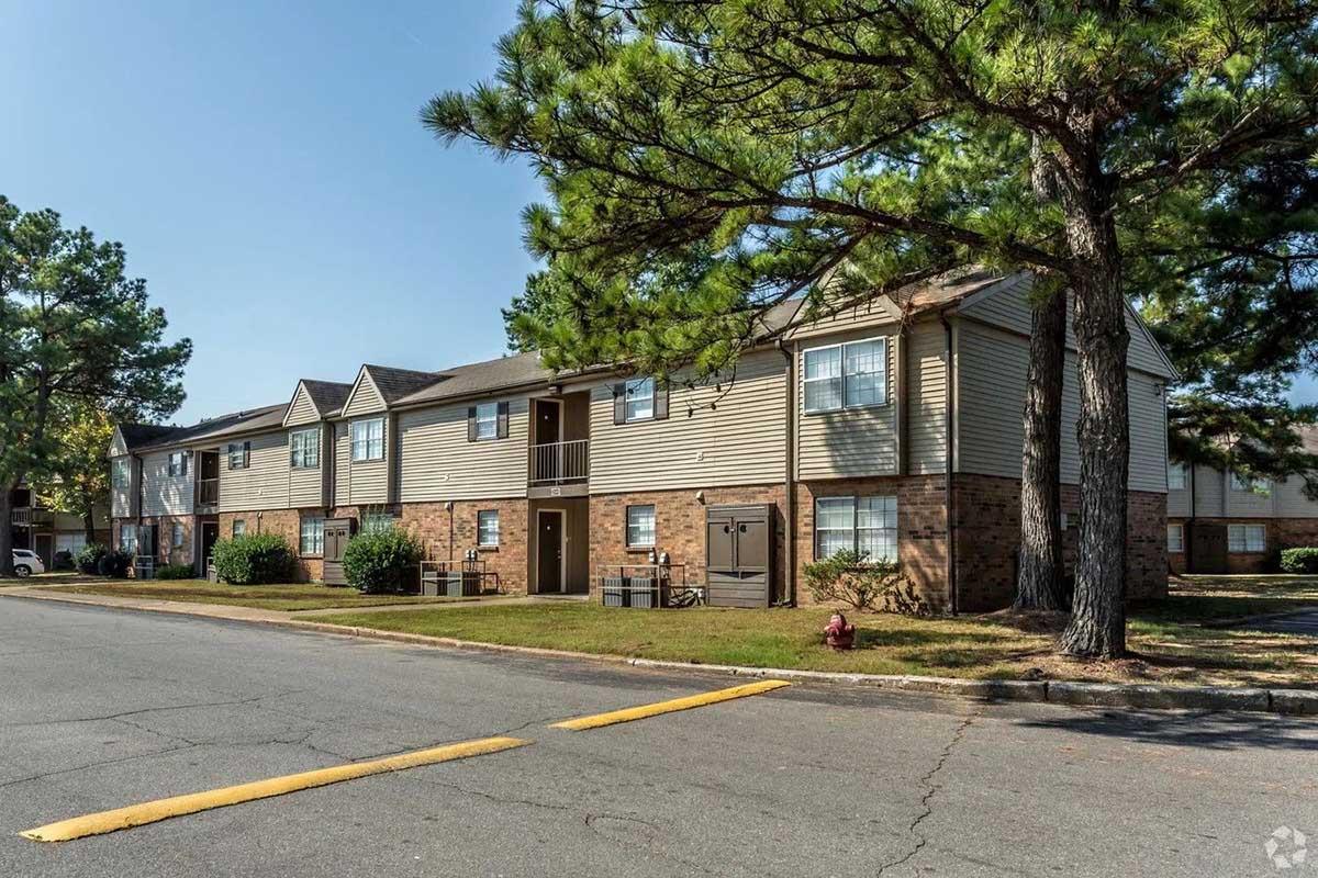 A well-maintained apartment complex featuring multiple two-story buildings. The buildings have light-colored siding with brick accents, surrounded by grassy areas and trees. The scene shows a clear blue sky and an asphalt road leading to the complex.