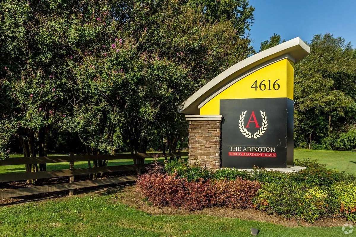 Sign for The Abington, an apartment complex, featuring the name and address "4616." The sign is a combination of yellow and stone elements, surrounded by landscaping with trees and bushes, set in a well-maintained outdoor area.
