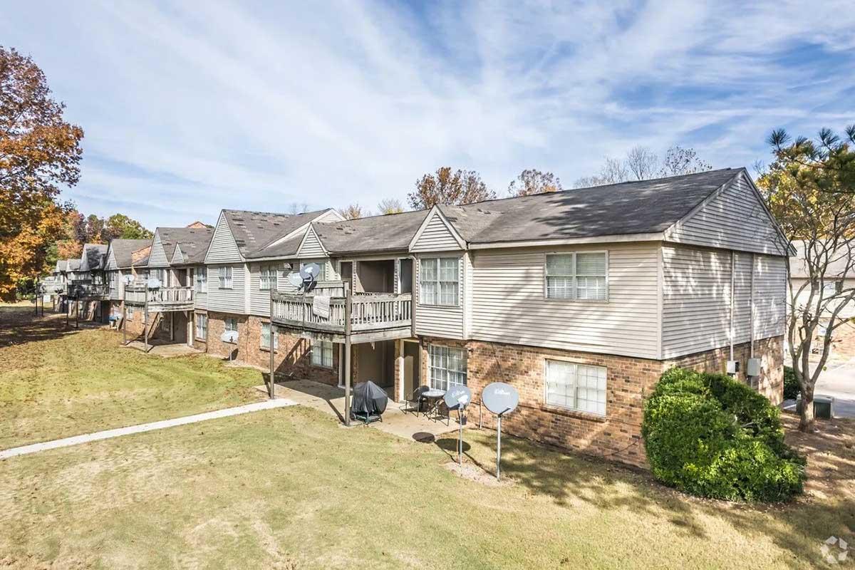 Two-story apartment building featuring multiple units with balconies. Surrounding grassy area with a few trees, and satellite dishes on the exterior. Clear blue sky with scattered clouds in the background, indicating a sunny day.
