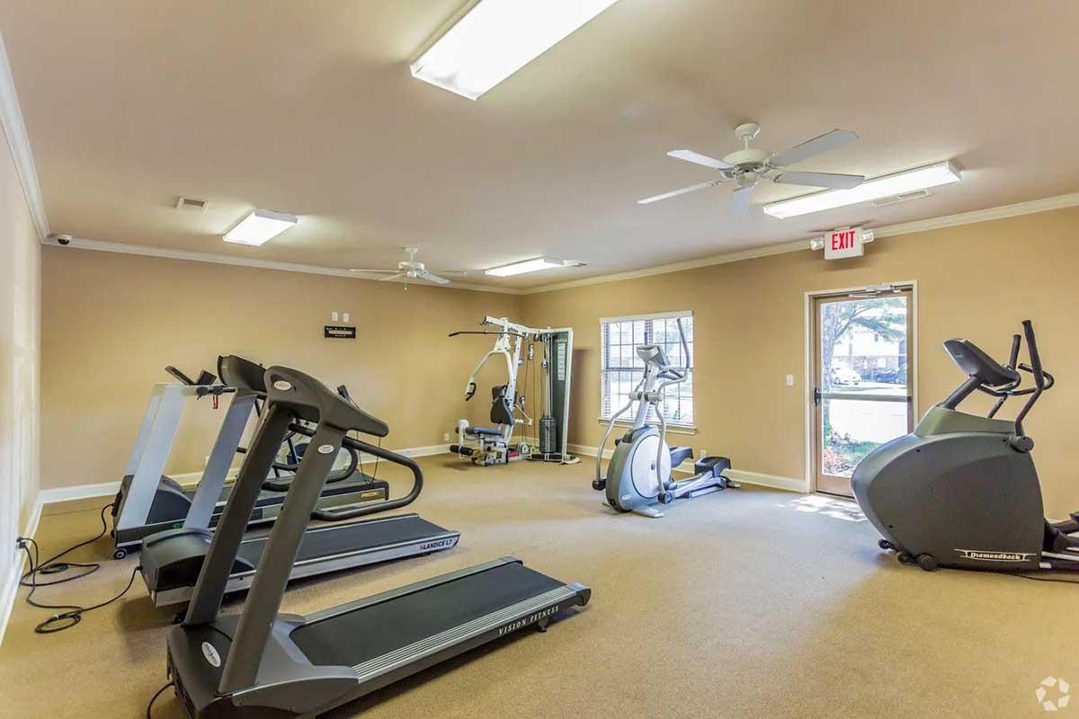 A well-lit gym featuring multiple treadmills, an elliptical machine, and strength training equipment. The room has light-colored walls, ceiling fans, and large windows allowing natural light to enter. The entrance door leads outside to a view of greenery.
