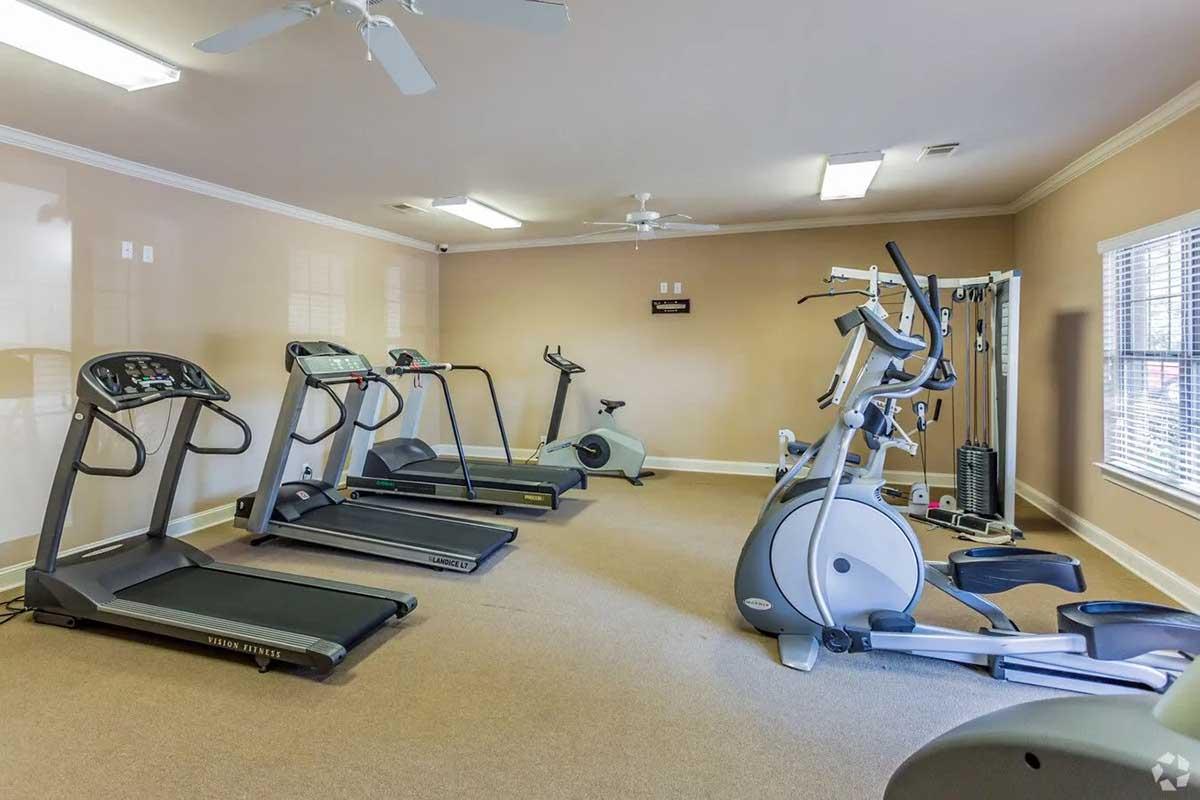 A fitness room featuring several exercise machines, including treadmills and an elliptical machine, arranged neatly on carpeted flooring. The space has beige walls and natural light coming through multiple windows, creating a bright and inviting atmosphere for workouts.