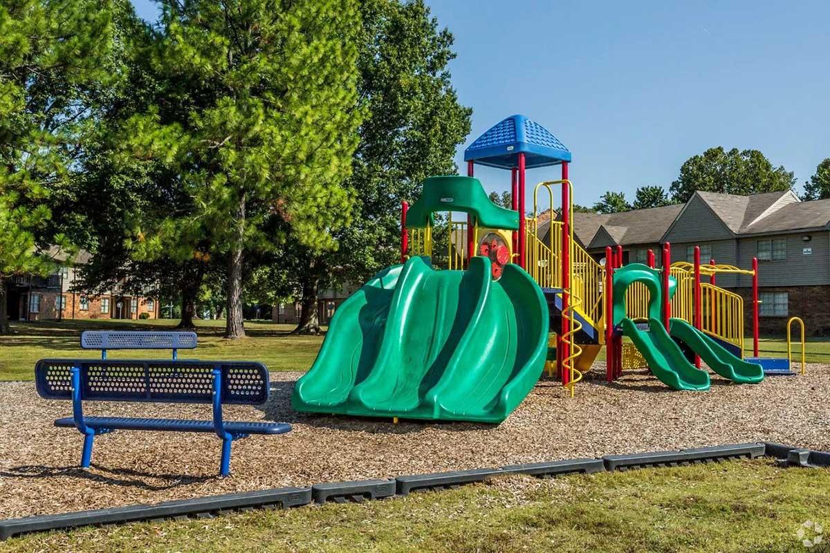 A colorful playground featuring multiple green slides, climbing structures, and a blue canopy, surrounded by trees and grass. There is a blue bench nearby, and residential buildings are visible in the background. The scene is bright and inviting, suitable for children to play.