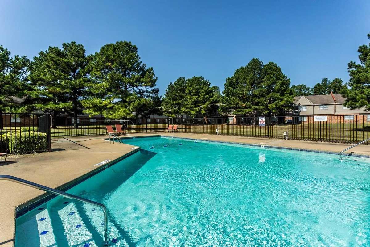 A clear blue swimming pool surrounded by a concrete deck and lounge chairs, with green trees in the background. The scene is bright and sunny, featuring a grassy area beyond the pool area with nearby buildings partially visible.