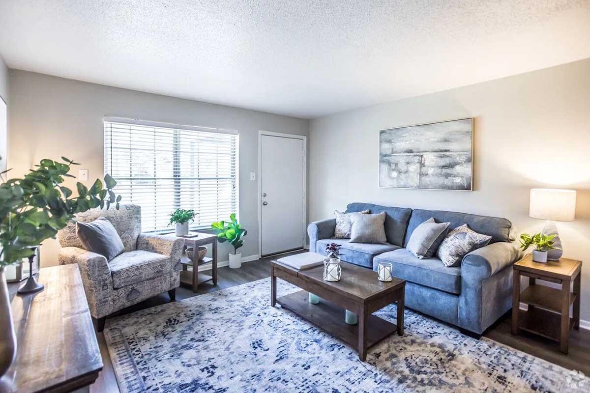 Cozy living room featuring a blue sofa and an accent chair with a patterned design. There's a wooden coffee table and side tables, with decorative plants and candles. A large window allows natural light, and a subtle area rug adds warmth to the space. A neutral artwork hangs above the sofa.