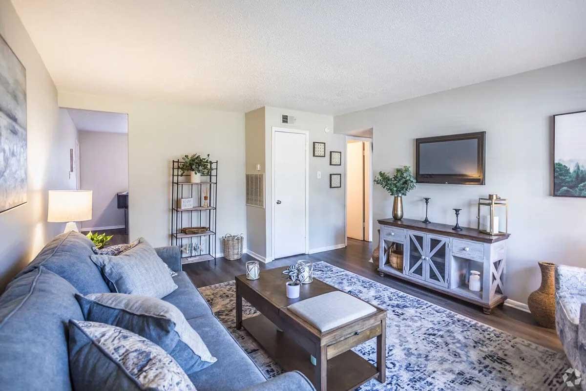 A cozy living room featuring a blue sofa with decorative pillows, a wooden coffee table, and a stylish sideboard. The walls are light-colored with artwork, and there's a lamp for warm lighting. A plant is visible in the corner, adding a touch of greenery. The space is well-lit and inviting.
