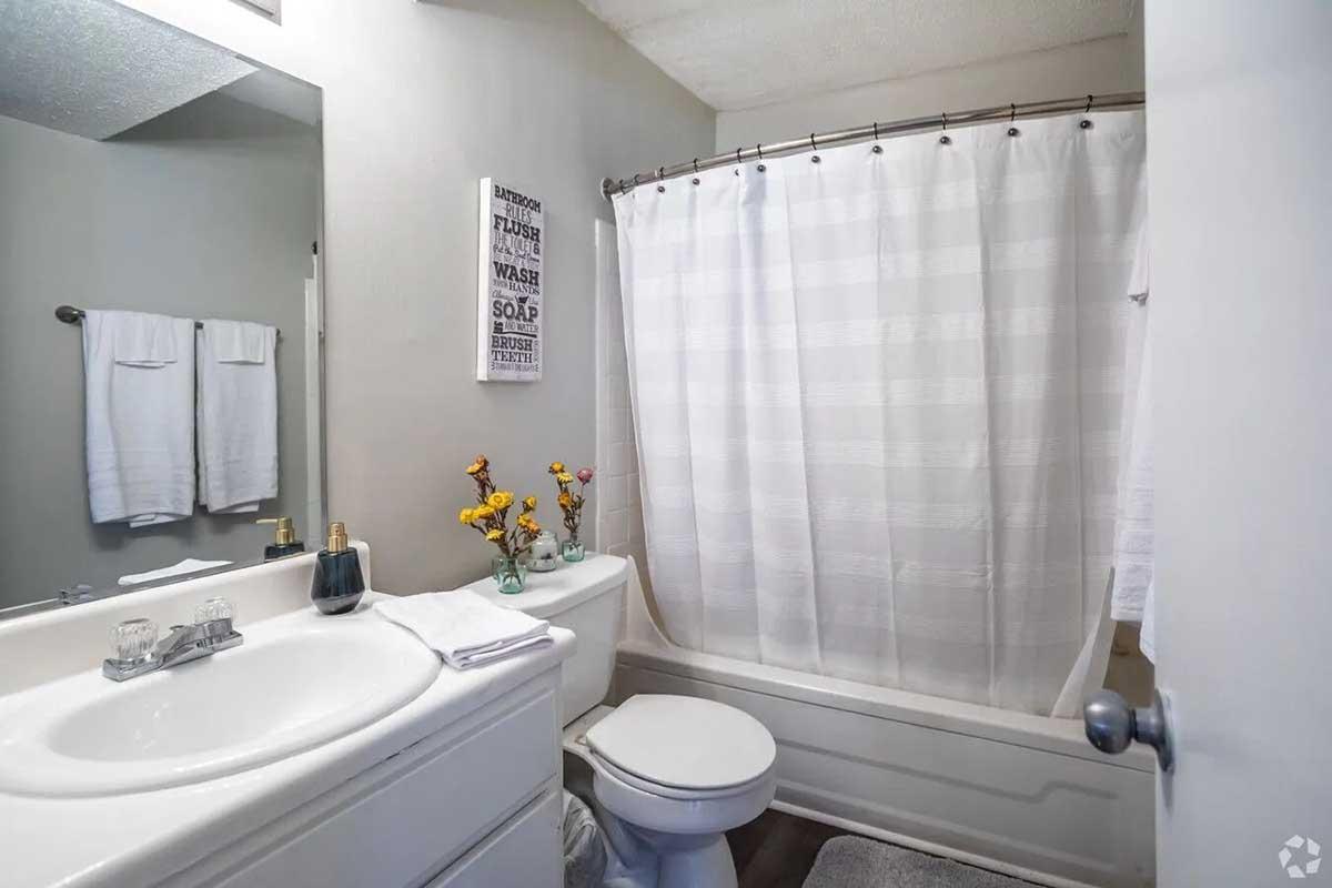 A clean, well-lit bathroom featuring a white bathtub with a shower curtain, a sink with a mirror above, and a toilet. White towels and a vase of flowers are neatly arranged on the countertop, adding a touch of decor to the space. A wall sign with bathroom-related text is visible.