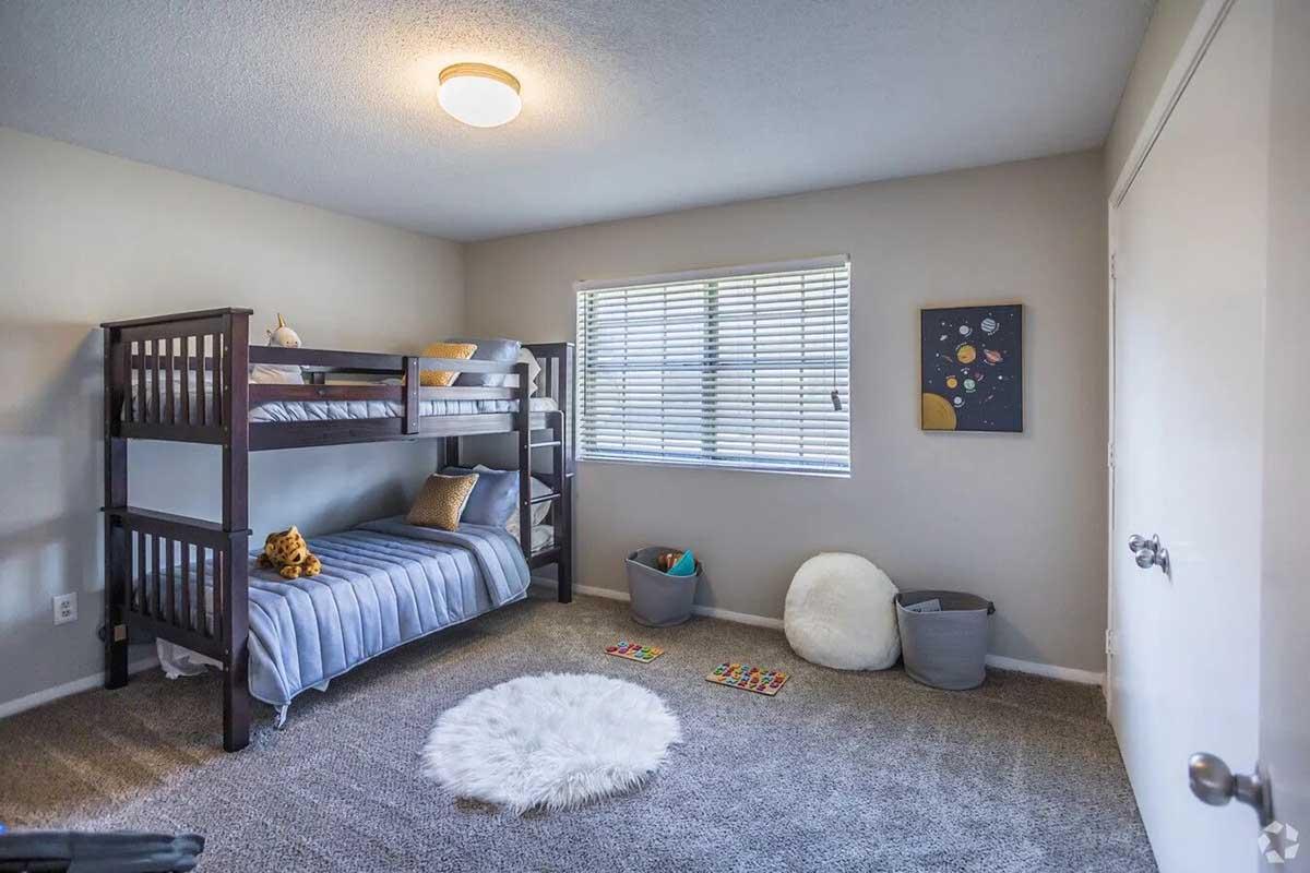 A cozy children's bedroom featuring a dark wood bunk bed with a blue and white blanket on the top bunk and a soft blue blanket on the bottom. Fluffy rugs on the carpeted floor, storage baskets, and a colorful space-themed wall art piece. Natural light streams through a window with white blinds.