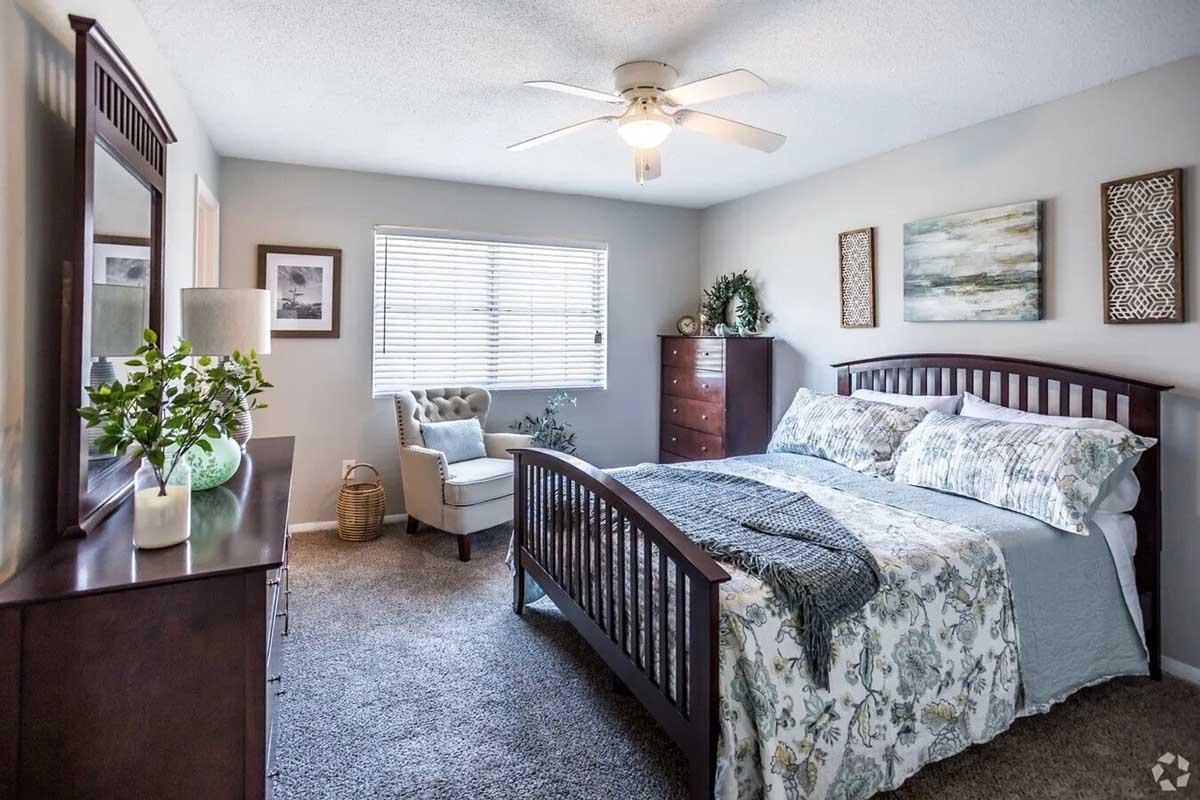 A well-lit bedroom featuring a cozy bed with decorative pillows and a soft blanket. There is a light-colored armchair in one corner, a wooden dresser, and a nightstand with a lamp. The walls are adorned with framed artwork, and a window allows natural light to fill the space. Soft, neutral colors create a serene atmosphere.