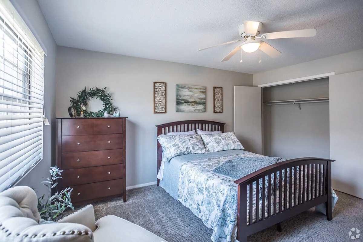 A well-lit bedroom featuring a wooden bed with blue and white bedding, a dark wood dresser, and two wall art pieces. A ceiling fan hangs above, and there’s a cozy chair in the corner. The room includes a closet with doors partially open and a window with blinds letting in natural light.