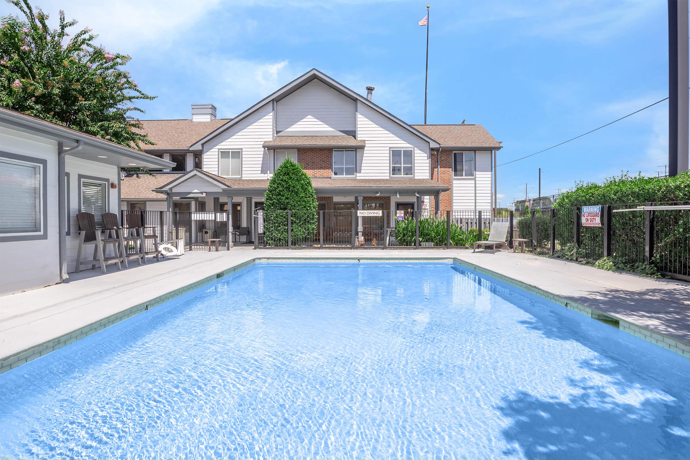 A clear swimming pool surrounded by a fenced area, with a residential building in the background. The building features multiple windows, and a flag can be seen on a pole. Lush greenery lines the pool area, and there are chairs positioned nearby for relaxation.