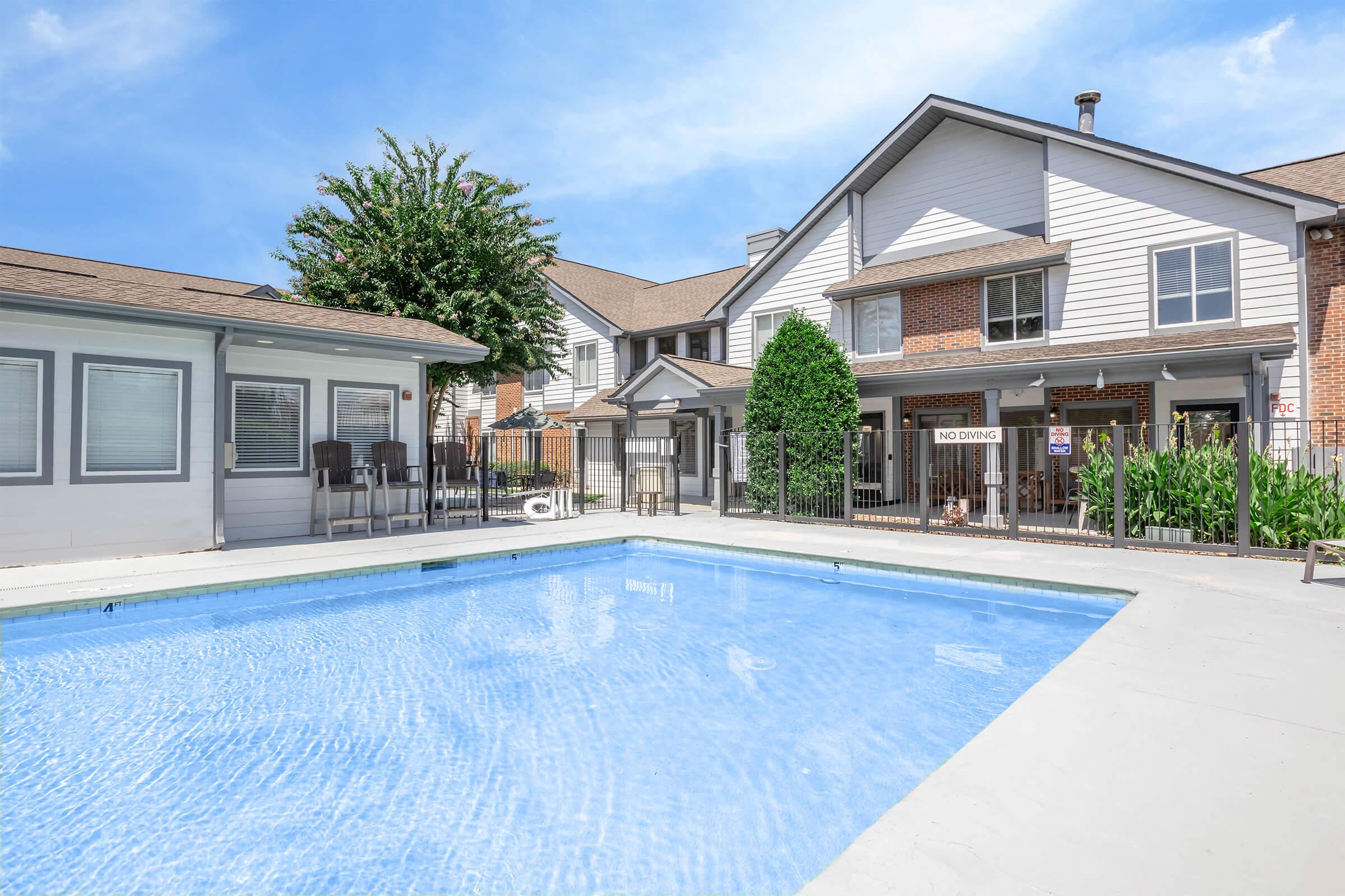 A bright outdoor swimming pool with clear blue water, surrounded by a well-maintained area featuring lounge chairs. In the background, there are residential buildings with a mix of brick and siding, and lush greenery. A "No Diving" sign is visible near the pool. The sky is clear and sunny.