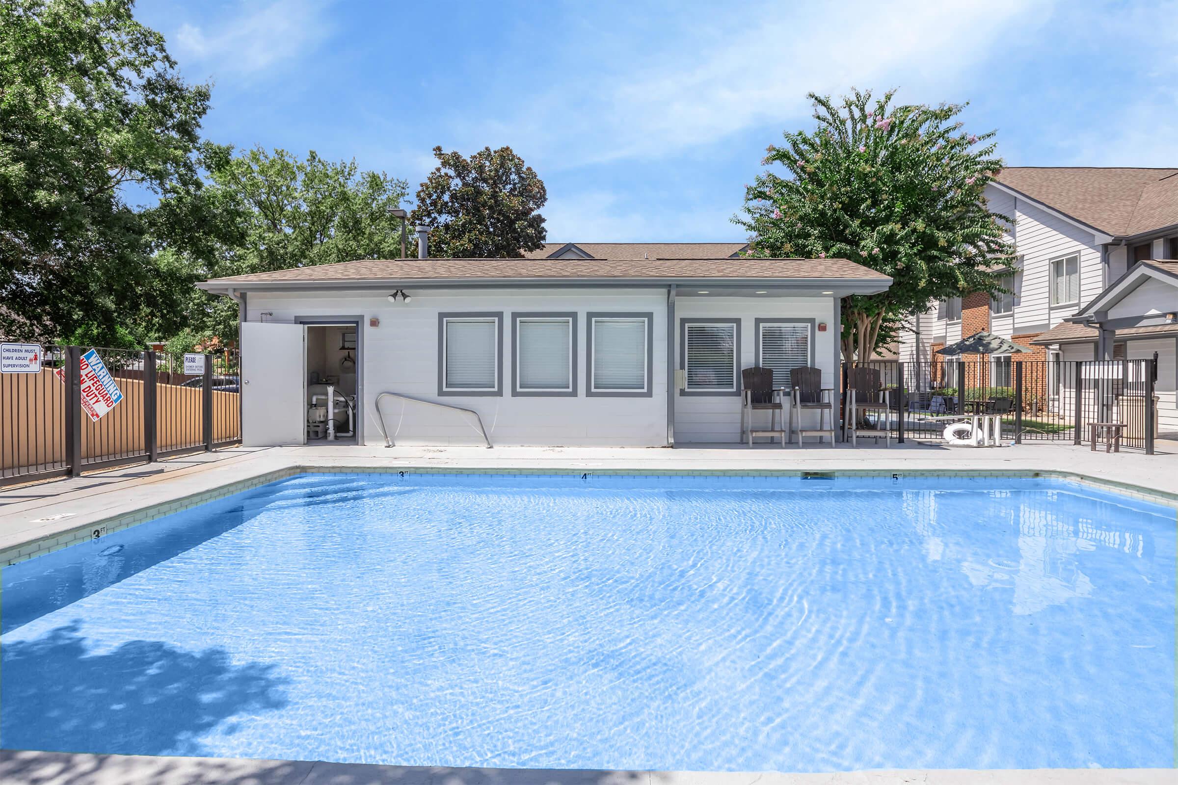 A clear blue swimming pool surrounded by a white fence and a small building with several windows. There are chairs and tables on a patio area beside the pool, along with trees and a residential building in the background. The sky is clear with a few clouds.