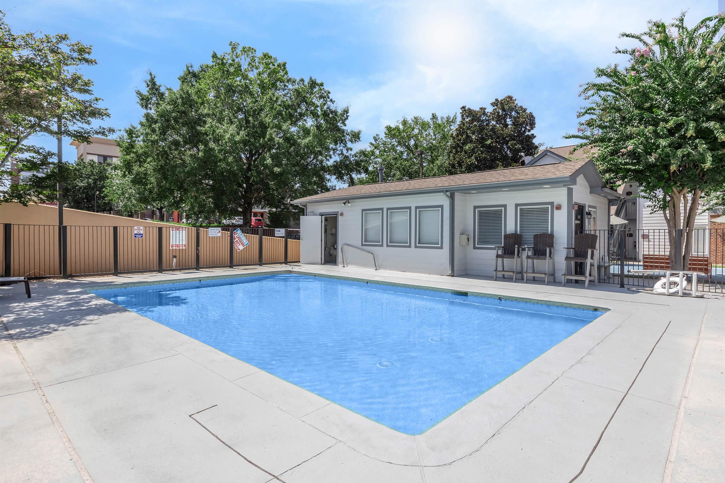A clean, outdoor swimming pool surrounded by a concrete deck and shaded by trees. Nearby, there's a modern building with large windows and seating areas. The sky is bright and blue, creating a refreshing atmosphere. Fencing surrounds the pool area for safety.
