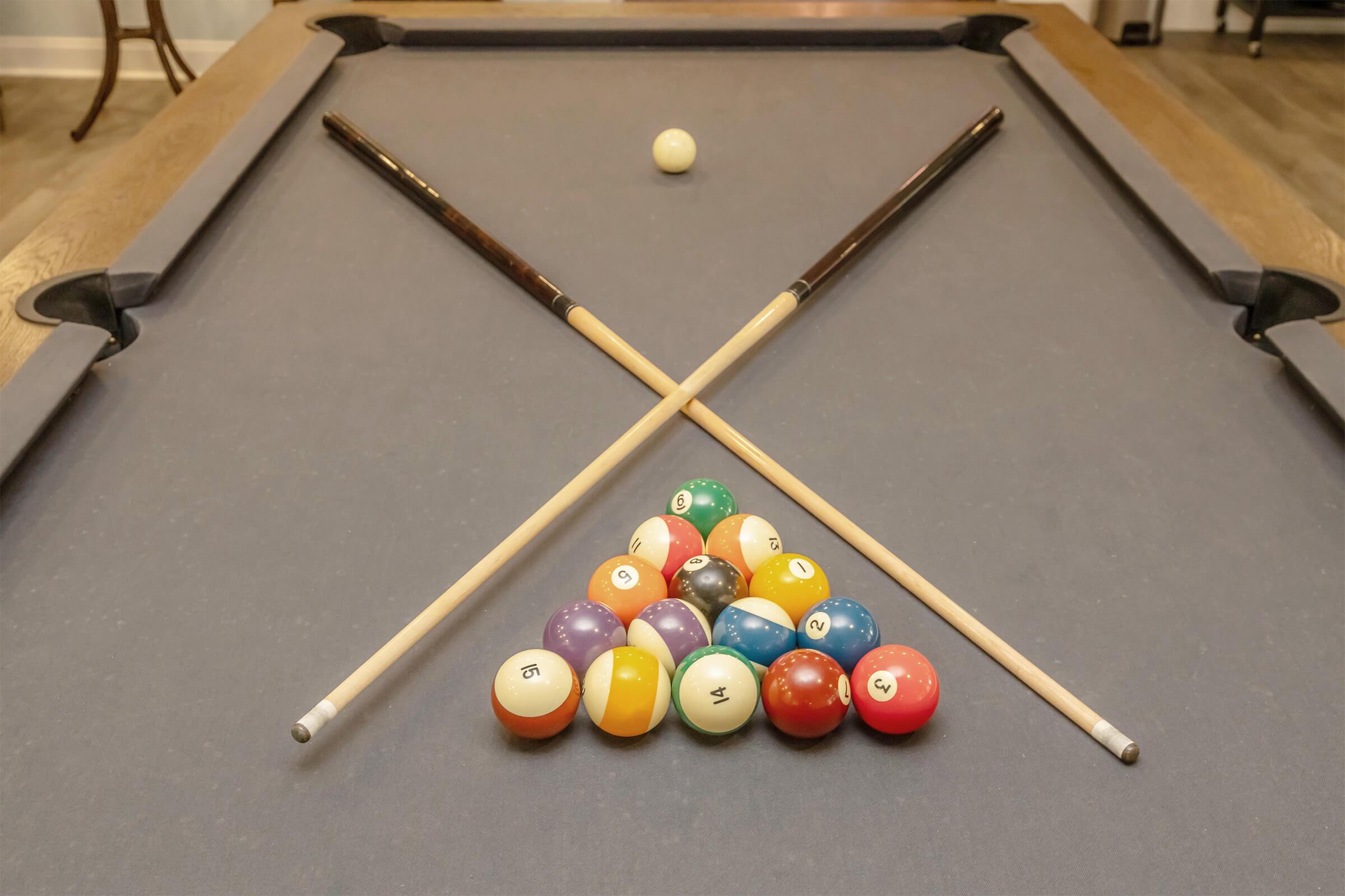 A pool table with a triangle rack of colored billiard balls arranged at the bottom. Two cue sticks are crossed over the rack, and a white cue ball is positioned at the top of the table. The table has a dark felt surface and wooden edges, creating a classic billiard setting.