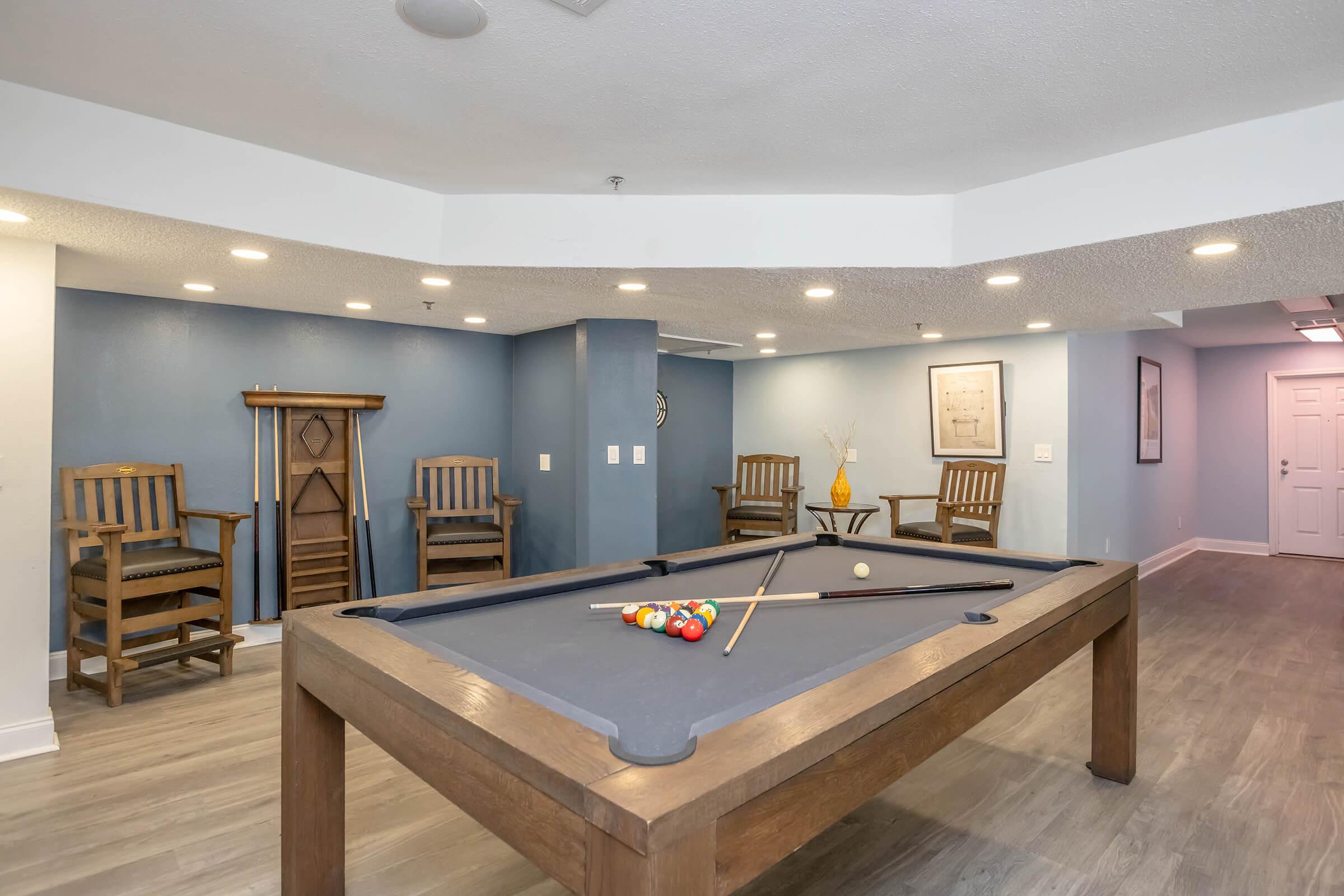 A spacious room featuring a dark pool table with colorful balls arranged on it. In the background, there are wooden chairs and a decorative wall. The lighting is soft, and the walls are painted in a calming blue tone, creating a relaxed atmosphere ideal for leisure activities.