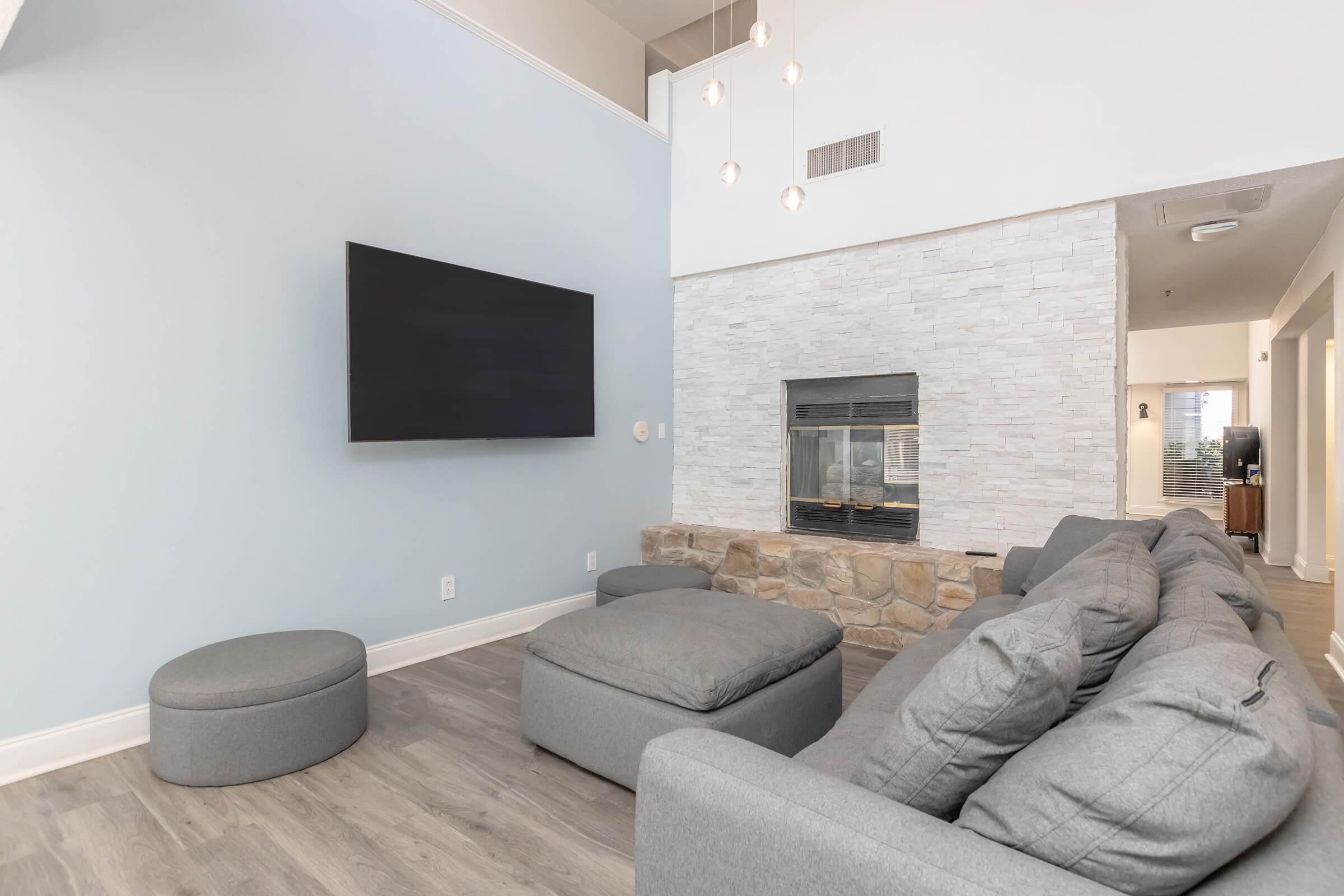 Modern living room featuring a wall-mounted flat-screen TV, a cozy stone fireplace, and a large gray sectional sofa with various cushions. A round ottoman complements the seating area. The walls are painted light blue, and the space is illuminated by stylish hanging lights.