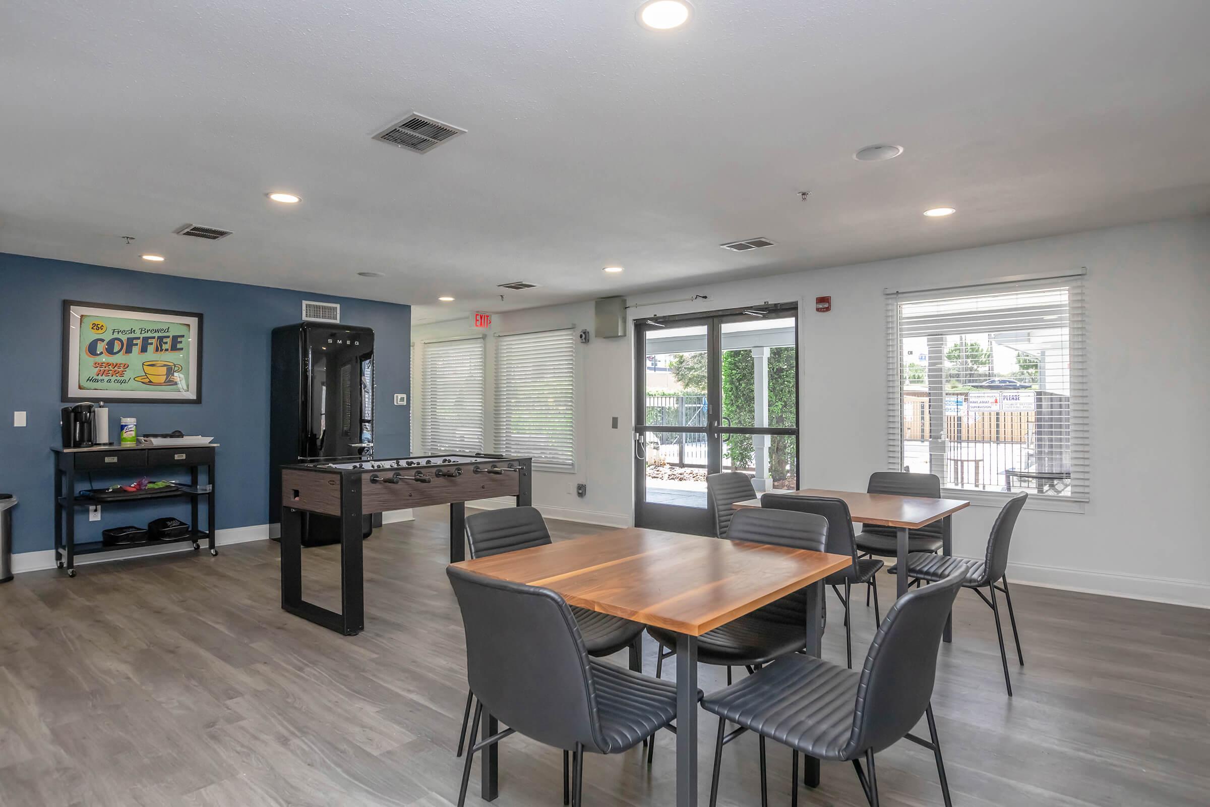 A modern lounge area featuring a large table with black chairs, a coffee station against a blue wall, and large windows allowing natural light. There’s a game table in the center, and the walls are painted in neutral tones, creating a welcoming atmosphere.