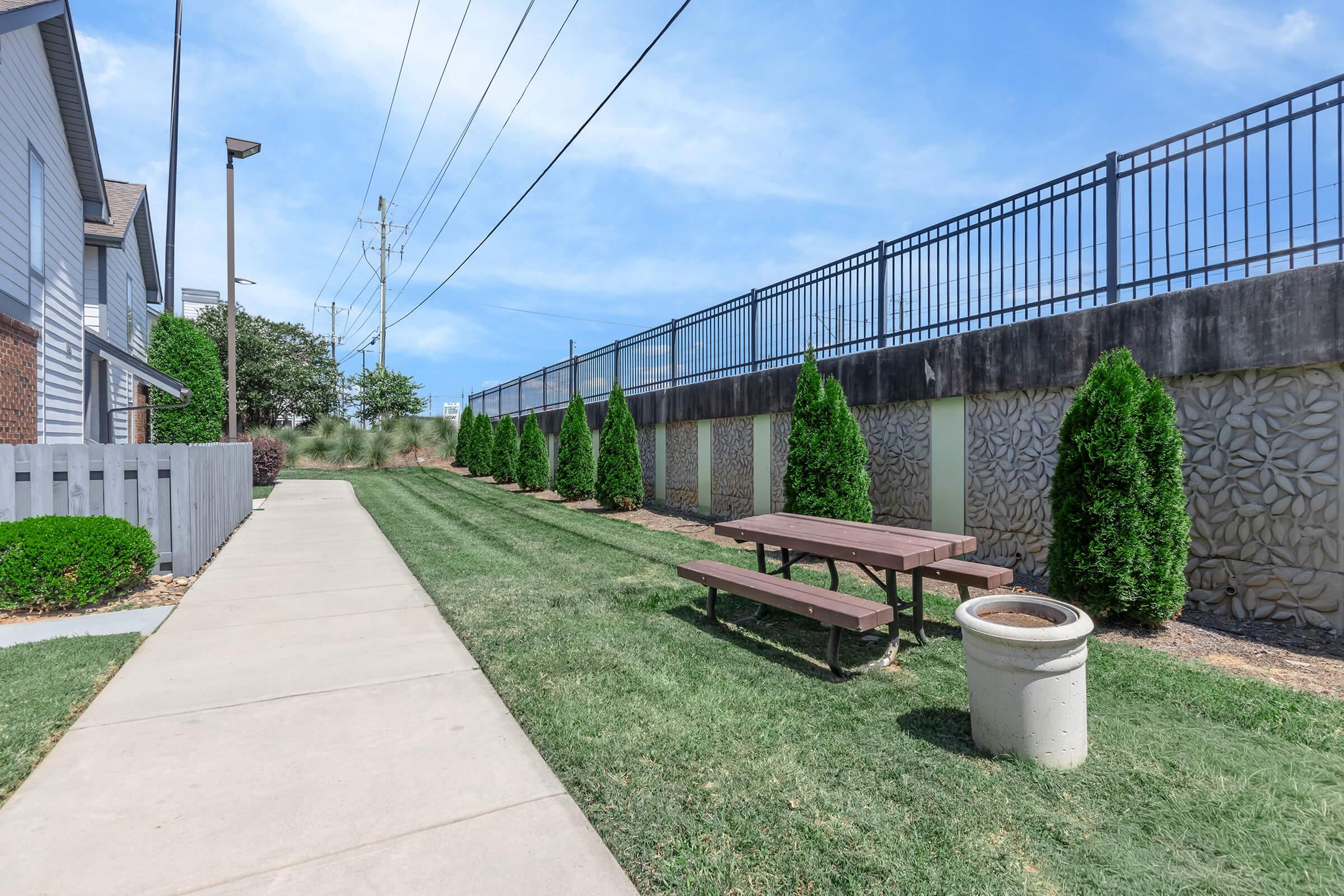 Pathway alongside a grassy area featuring a picnic table and trash can, bordered by small shrubs and trees. A tall fence runs parallel to the path, with power lines visible in the background under a clear blue sky.