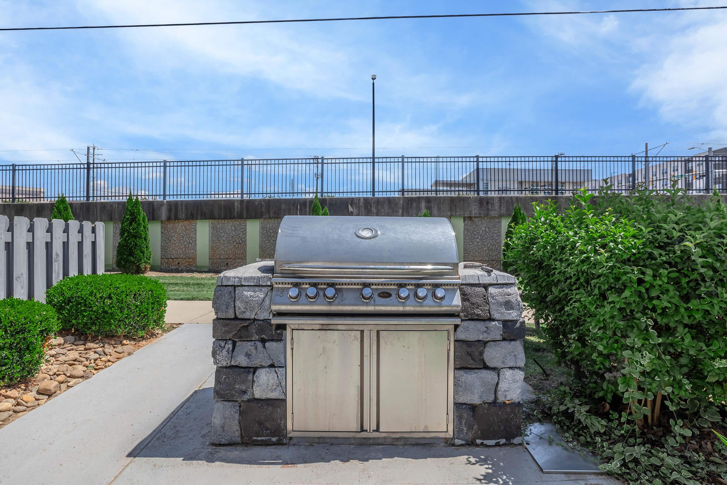 A stainless steel outdoor grill set on a stone base, surrounded by a concrete path, green bushes, and small decorative rocks. There’s a fence and a railing in the background, under a blue sky with scattered clouds.