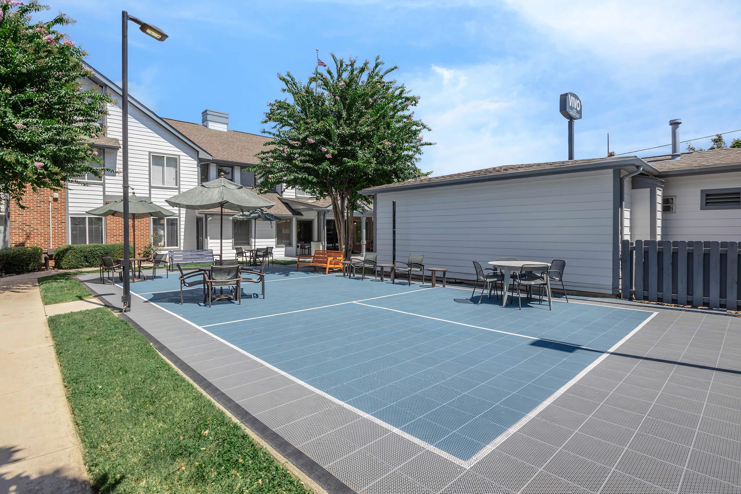 Outdoor recreational area with a gray tiled surface, featuring several tables and chairs, umbrellas for shade, and surrounding greenery. In the background, there are residential buildings and a sign partially visible. The setting is bright and inviting, ideal for relaxation or socializing.