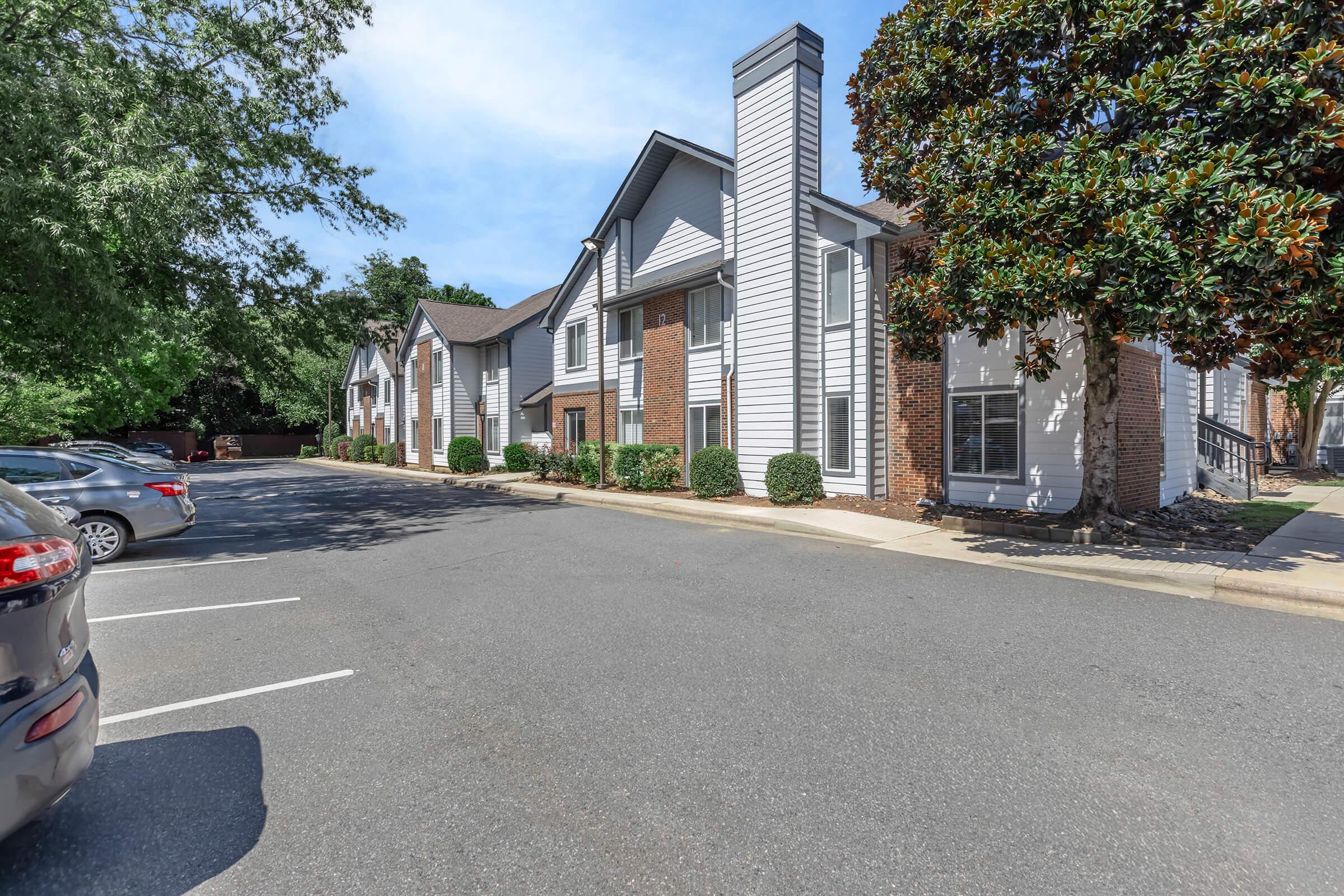 A residential apartment complex featuring two-story buildings with a mix of brick and white siding. There are several parked cars in the foreground and well-maintained landscaping with shrubs and trees surrounding the entrance. The sky is clear and blue, indicating a sunny day.