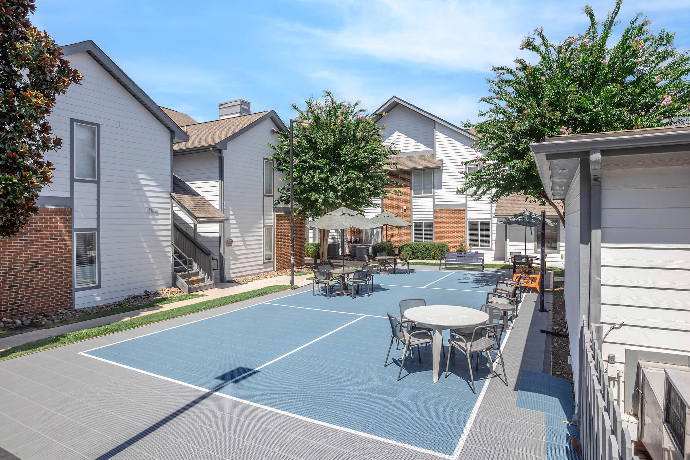 A spacious outdoor courtyard featuring blue tiled flooring, surrounded by landscaped greenery and trees. There are multiple seating areas with tables and chairs, including a shaded umbrella area. Nearby buildings have a modern architectural design with a mix of brick and siding.