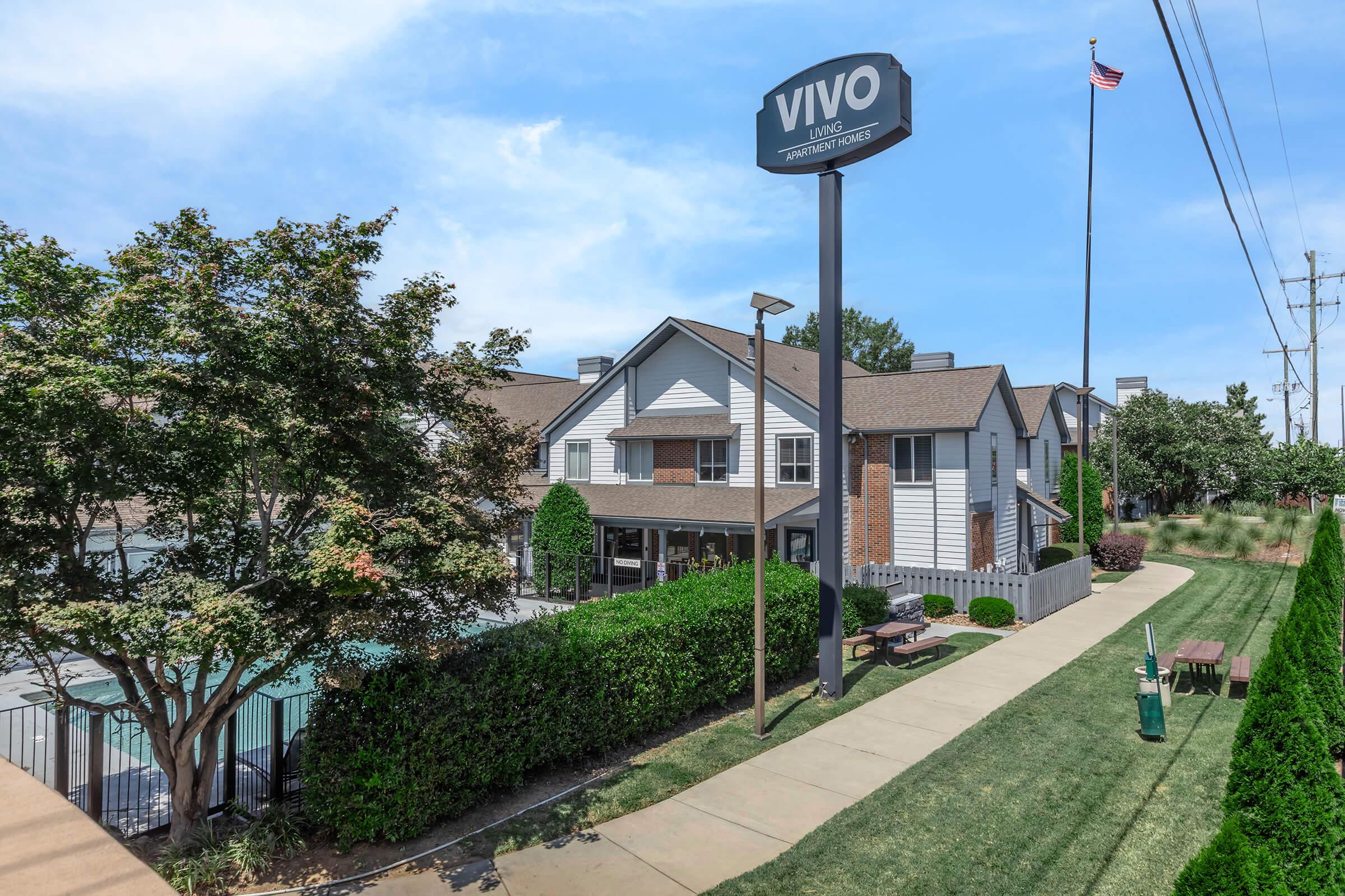 Exterior view of Vivo Living Apartments, featuring a two-story building with a pool visible, surrounded by well-maintained grass and shrubs. A sign with the "VIVO" logo is prominently displayed, alongside an American flag. Clear blue sky enhances the pleasant outdoor setting.