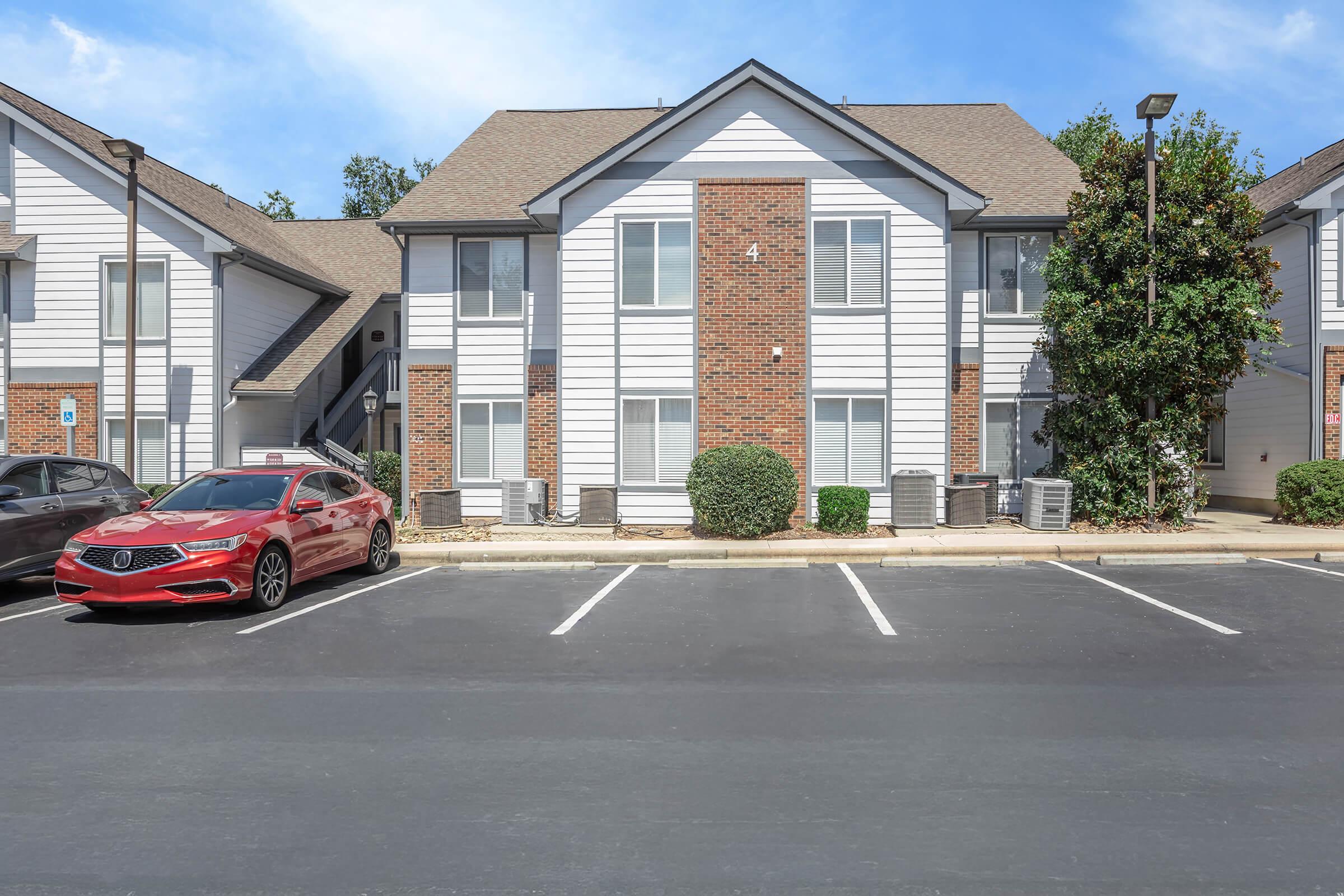 Exterior view of a two-story apartment building with a brick and white siding façade. There are two cars parked in front, one red sedan and one gray vehicle. The parking area is well-maintained, with shrubs and trees surrounding the building, and a clear blue sky above.