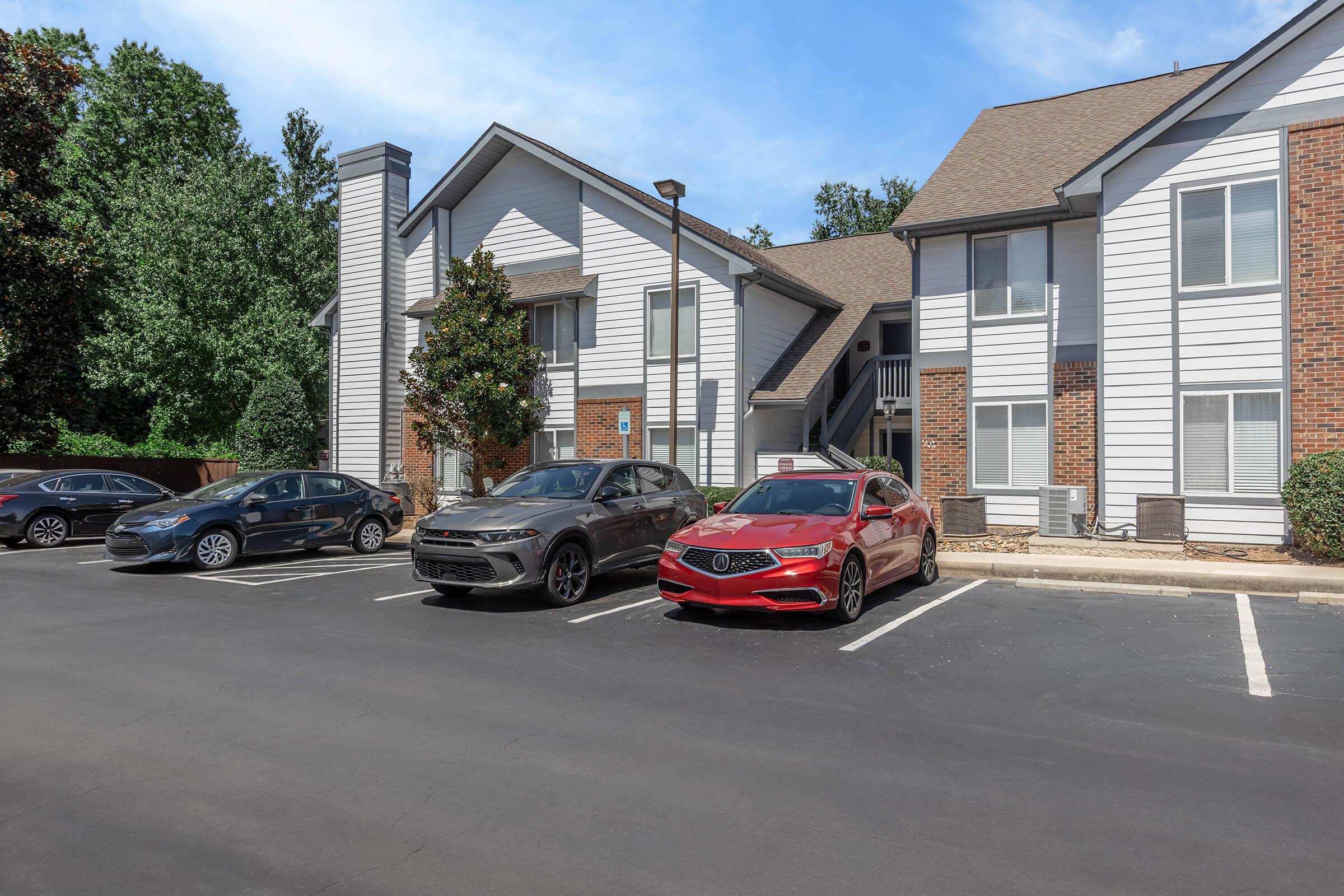 A parking area in front of a multi-story residential building. Two cars, one red and one gray, are parked in the foreground, along with another vehicle in the distance. The building features a mix of brick and siding, surrounded by trees and blue sky in the background.