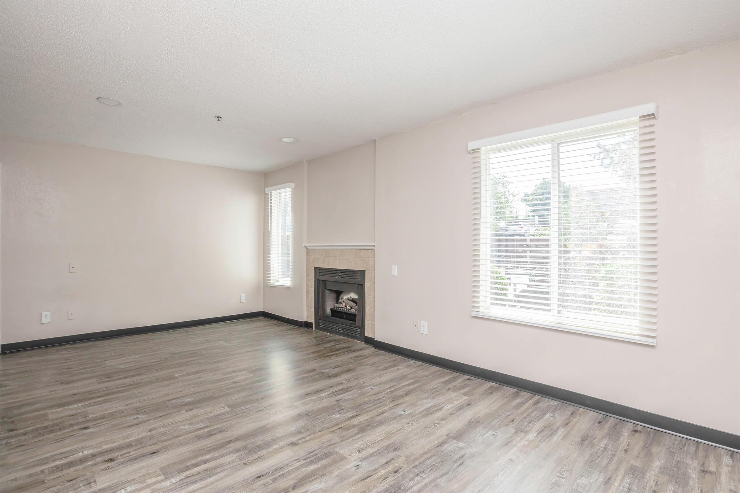 A sunlit living room featuring light walls and a fireplace. Large windows allow natural light to flood the space, and the floor is covered with modern, light-colored laminate. The room is empty, providing a blank canvas for decoration.