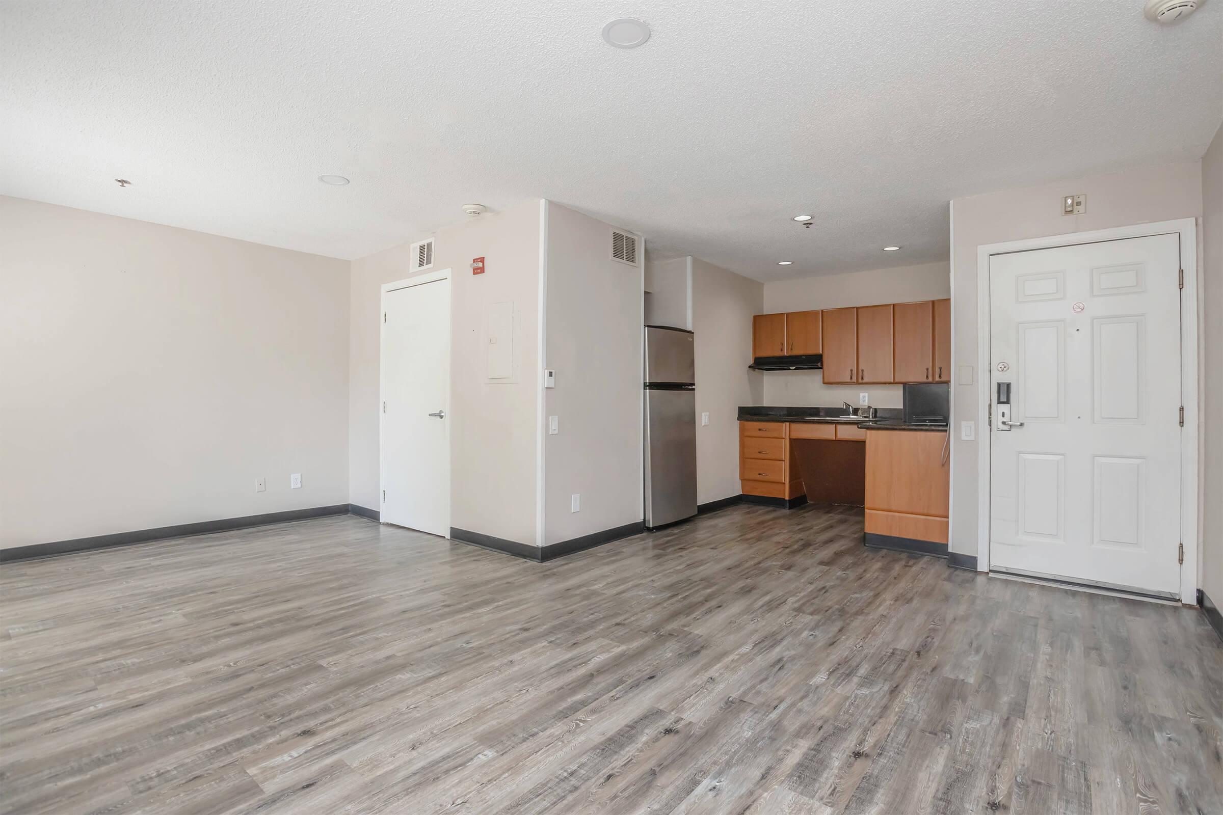 A spacious, modern studio apartment featuring light-colored walls, gray laminate flooring, and an open layout. The kitchenette is equipped with wooden cabinets, a refrigerator, and a stove. A front door with a small window is visible, and the room has natural light from overhead fixtures.