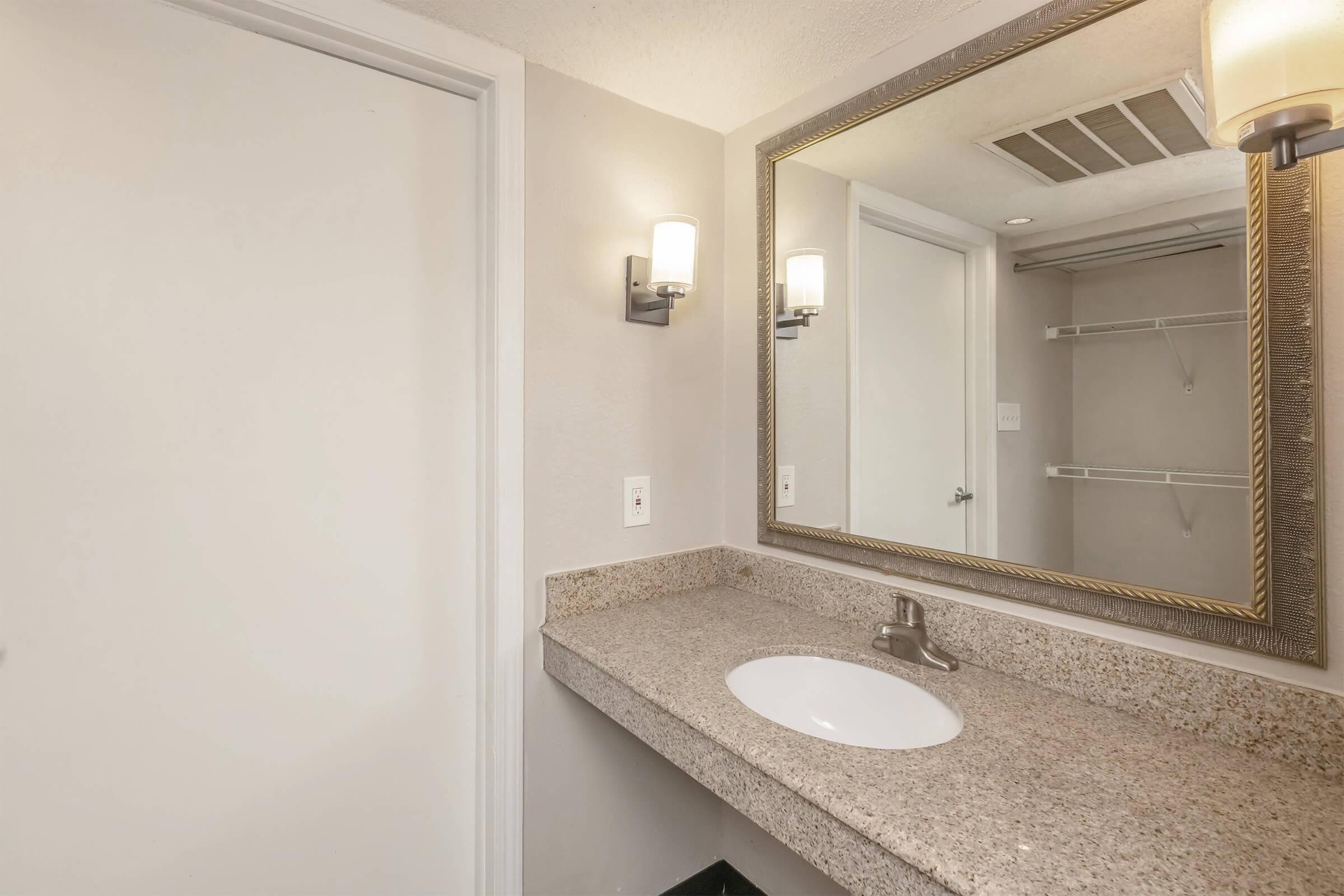 A clean and modern bathroom with a countertop, sink, and mirror. The walls are light grey, and there is a wall-mounted light fixture next to the mirror. A closet space is visible in the rear, with a simple door to the left. The overall design is functional and well-lit.