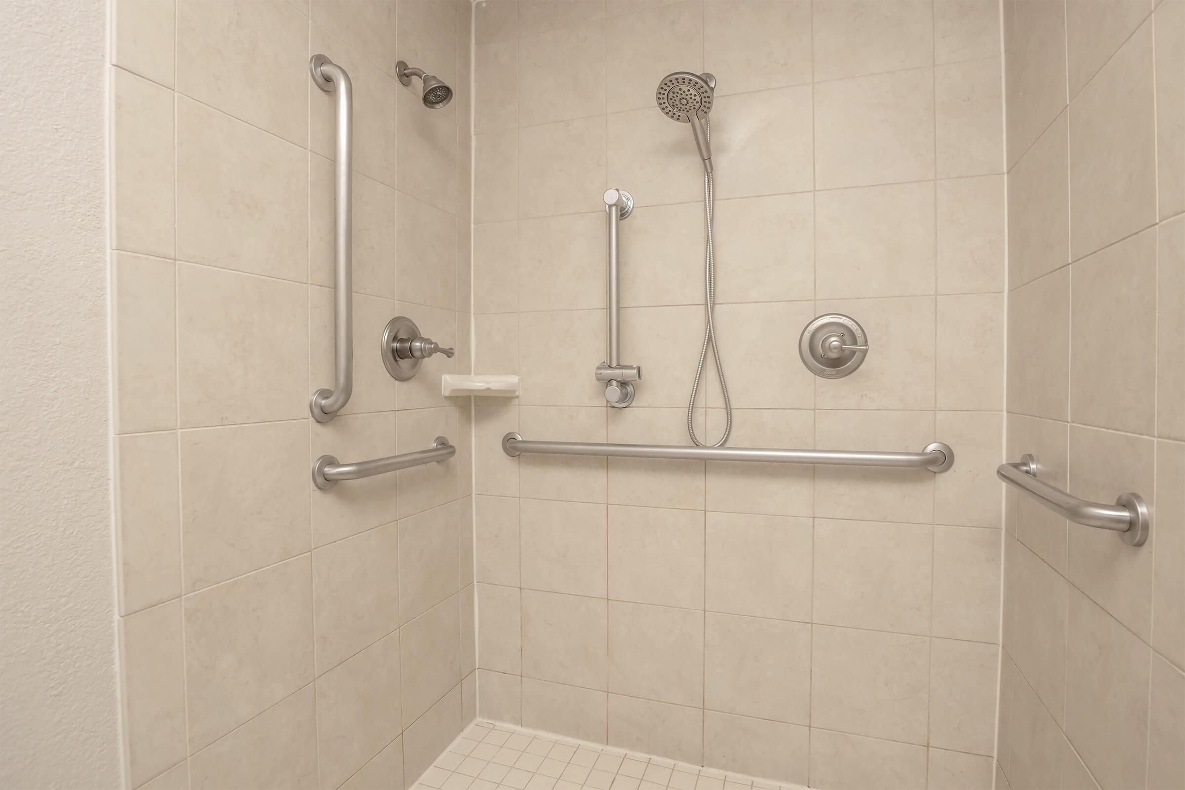 A clean, well-lit shower stall featuring light-colored tiled walls and a non-slip floor. It includes grab bars for safety, a handheld showerhead, and a wall-mounted shower control, designed for accessibility. The overall atmosphere is functional and modern.