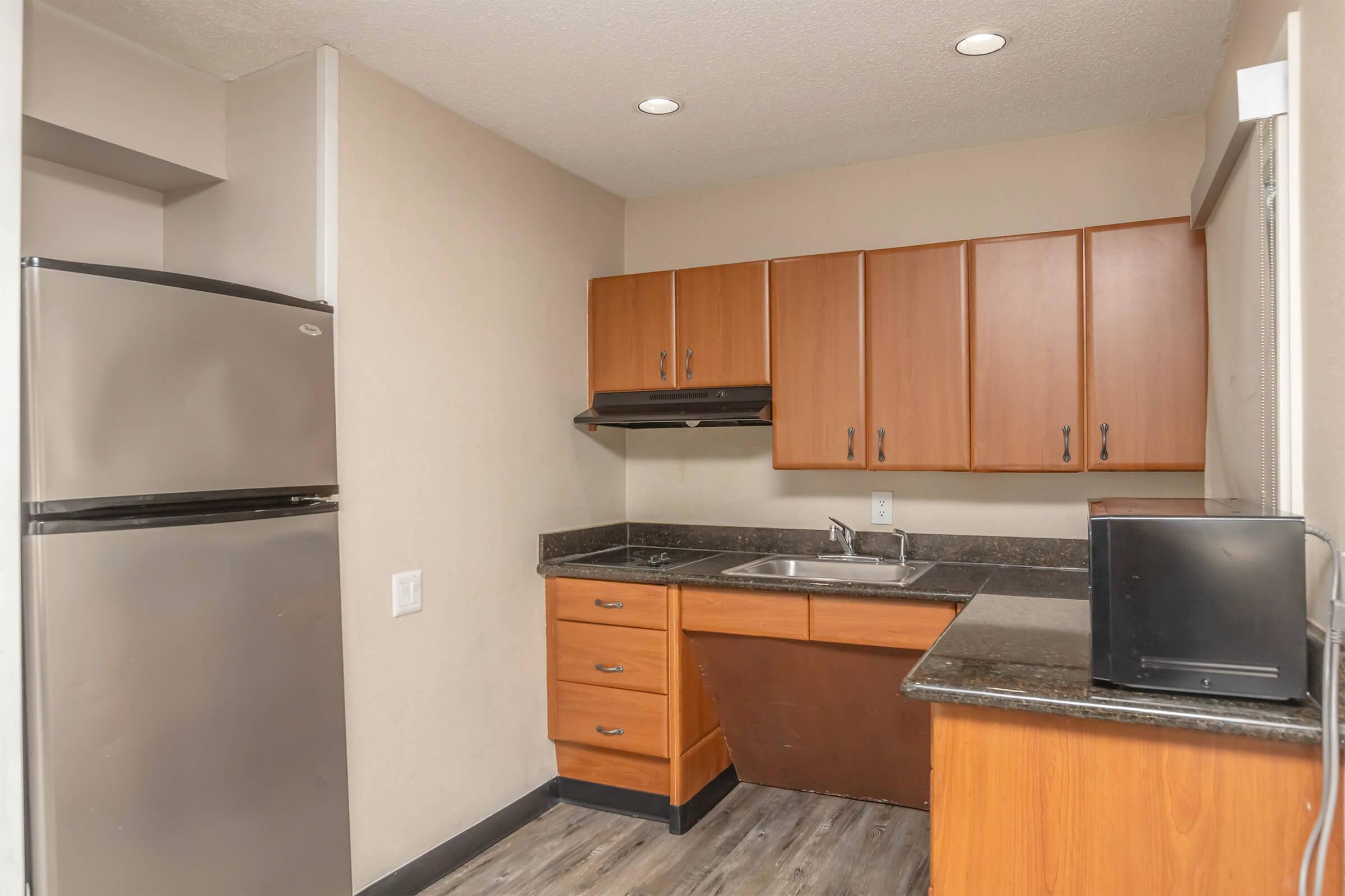 Modern kitchen featuring a stainless steel refrigerator, wooden cabinets, black countertop, and a sink. A microwave is also present on the counter. The walls are a neutral beige, and the flooring is dark wood-like laminate. Bright overhead lighting illuminates the space.