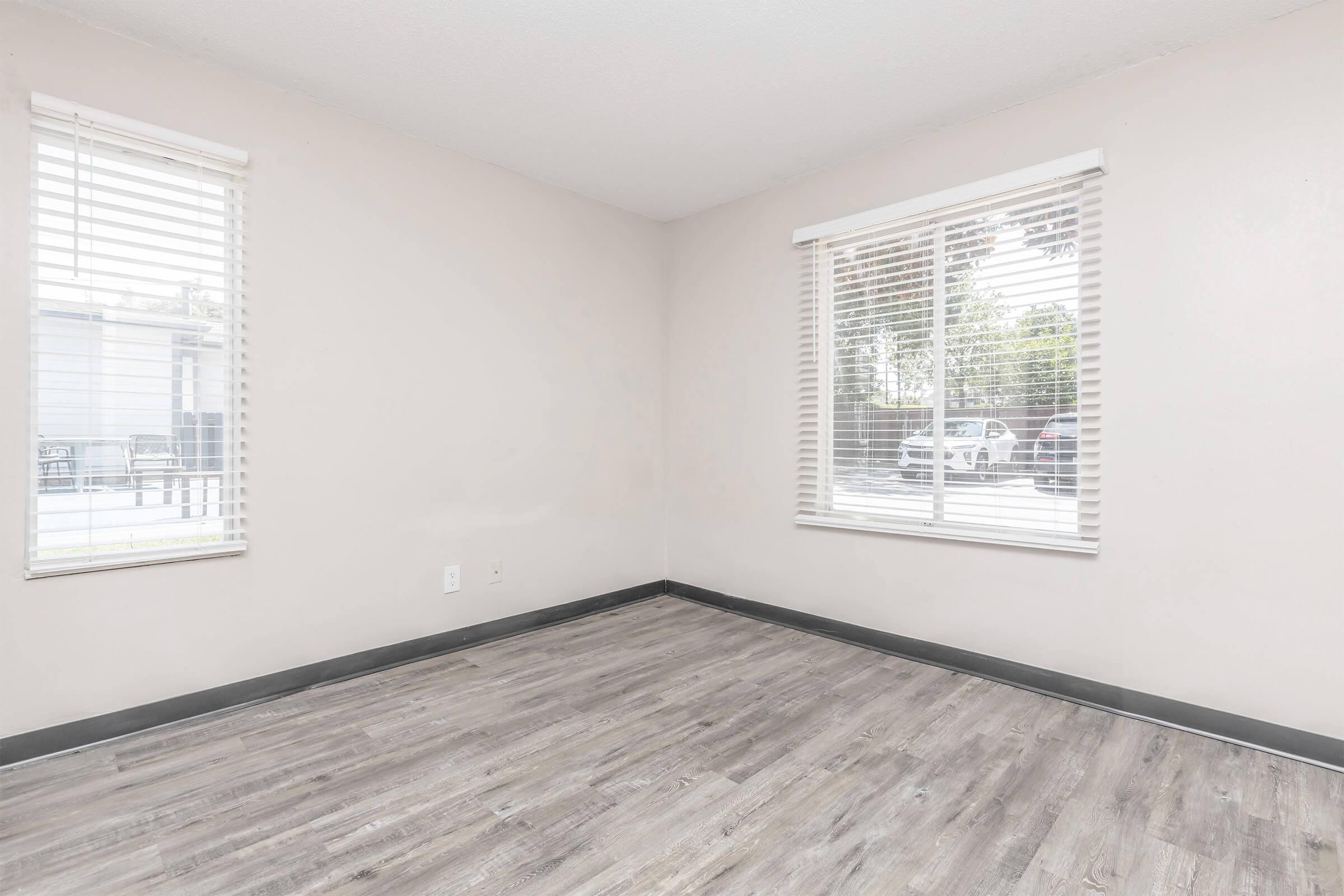 Empty room with light-colored walls and wooden flooring. There are two windows with blinds allowing natural light to filter in, and a view outside. The room is unfurnished, giving a spacious and airy feel.