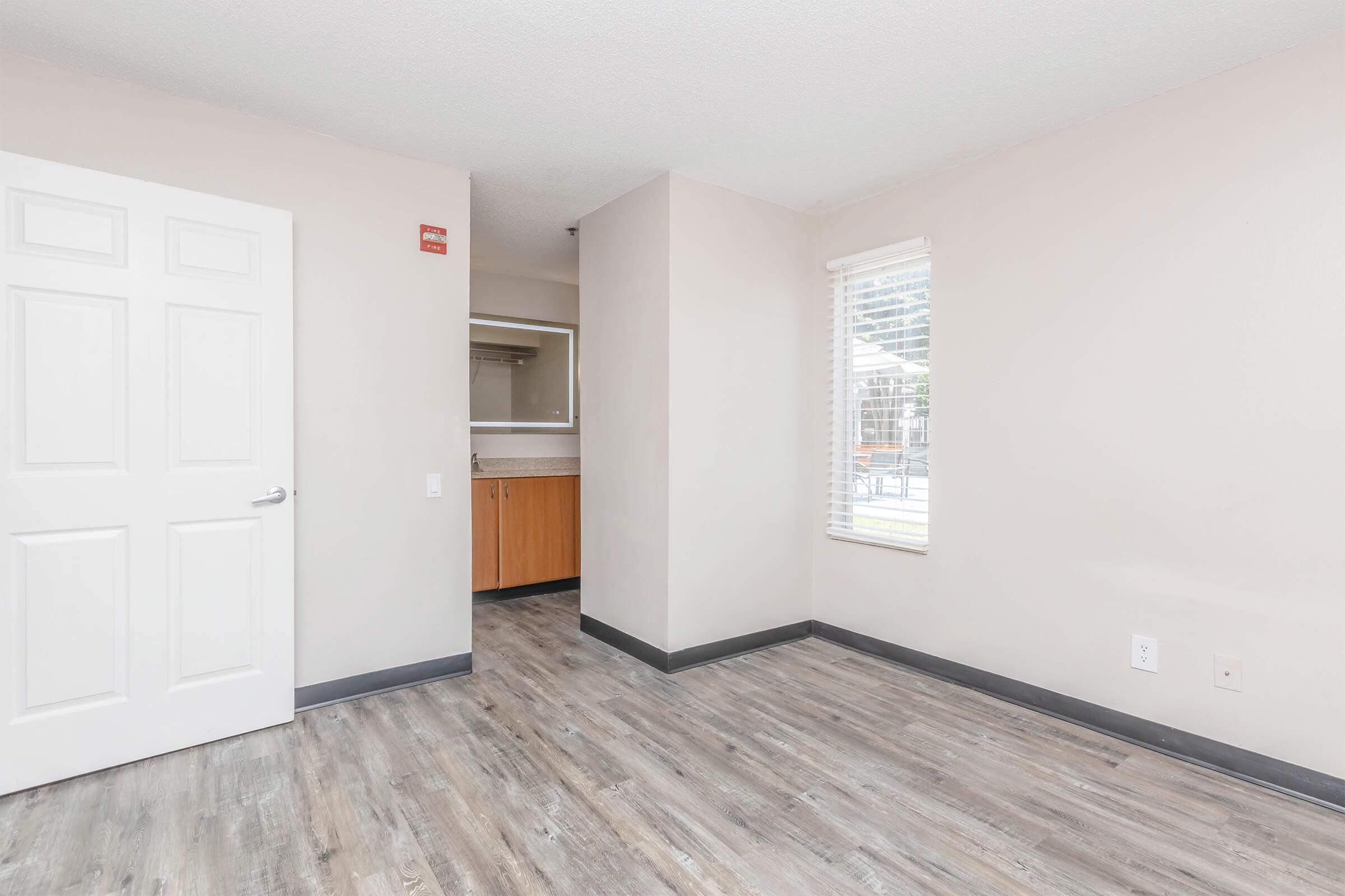 Empty room with light-colored walls, a white door, and a large window. The flooring is wood-like, and there are kitchen cabinets visible in the background. Natural light illuminates the space, giving it a clean and open feel.