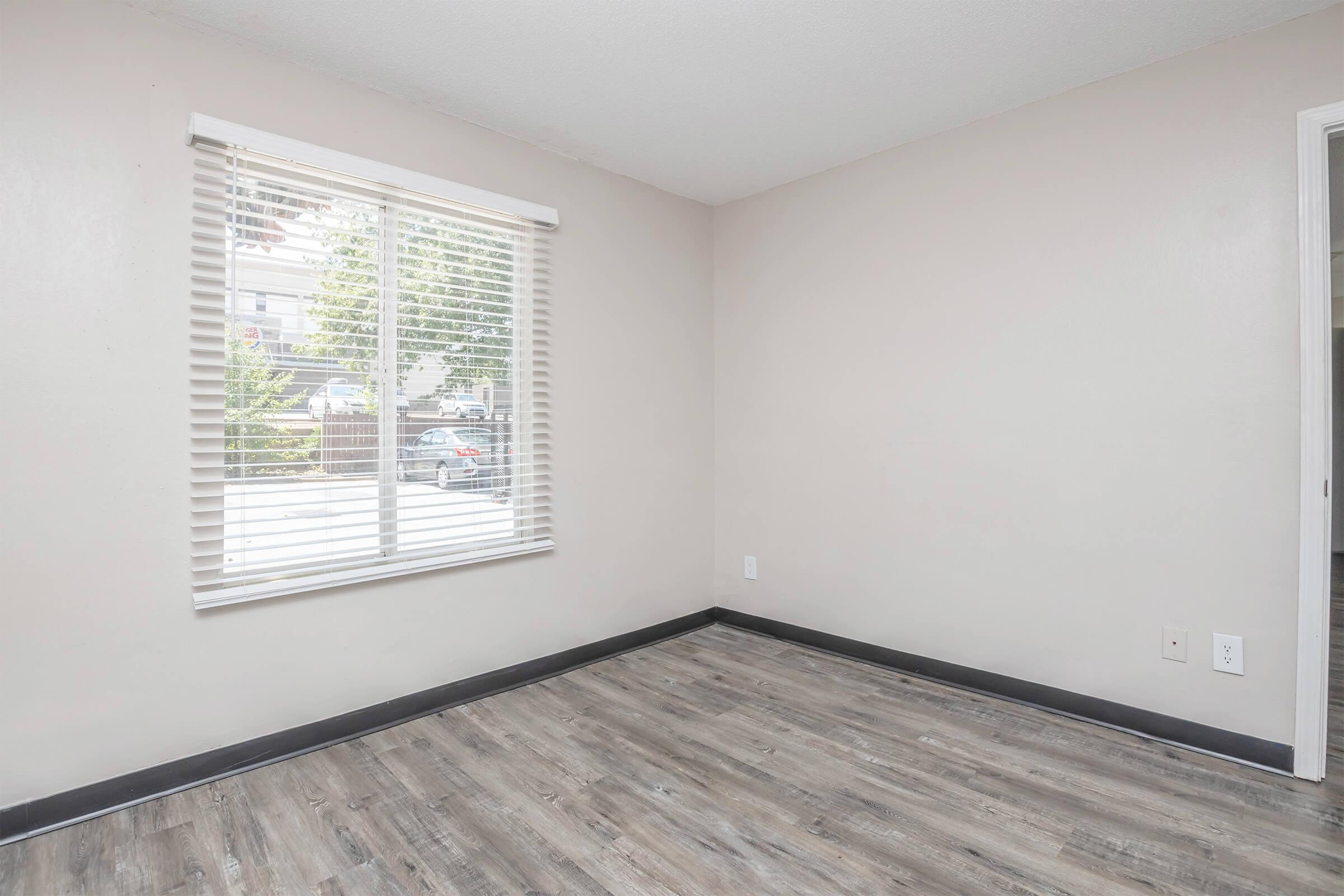 Empty room with light-colored walls and a faux wood floor. A window with white blinds lets in natural light. There are no furnishings, and the space appears clean and ready for moving in. A door is visible on the right side of the image, leading to another area.