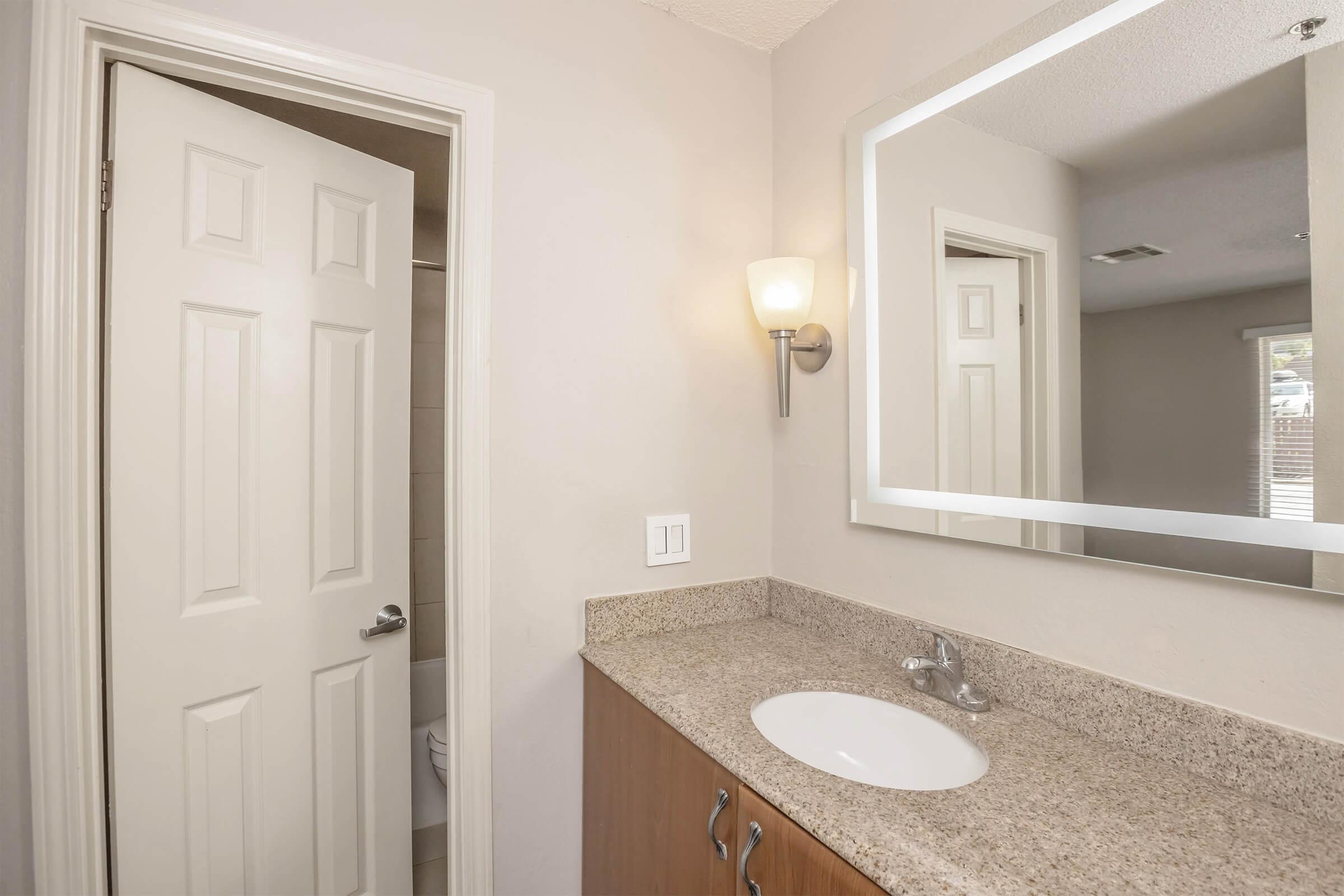 A bathroom interior featuring a light-colored countertop with a round sink, a wall-mounted mirror, and a wall sconce. A partially open door leads to a shower area, while the rest of the room appears neutral and well-lit, showcasing a clean and modern design.