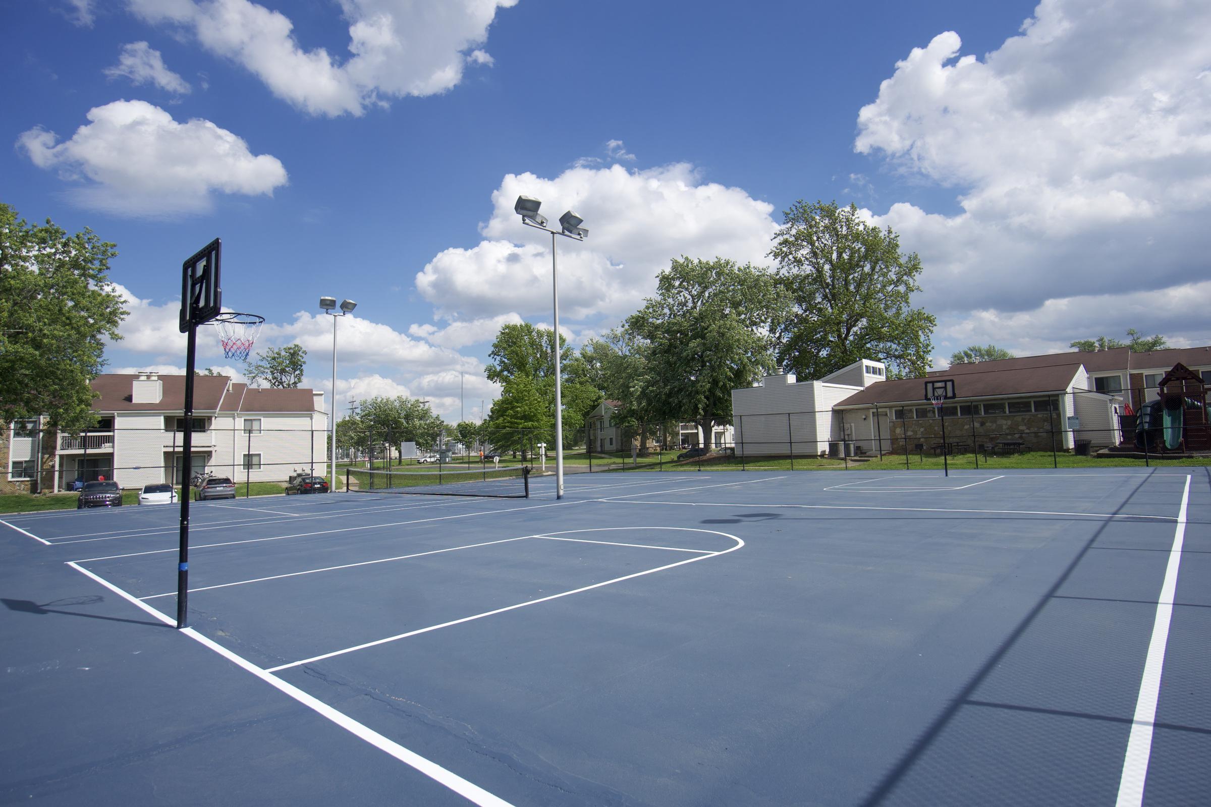 A view of a basketball court under a sunny blue sky with fluffy clouds. The court has two basketball hoops on opposite ends and is surrounded by greenery and residential buildings in the background. It's a clear day, and the court looks newly painted, inviting for players.